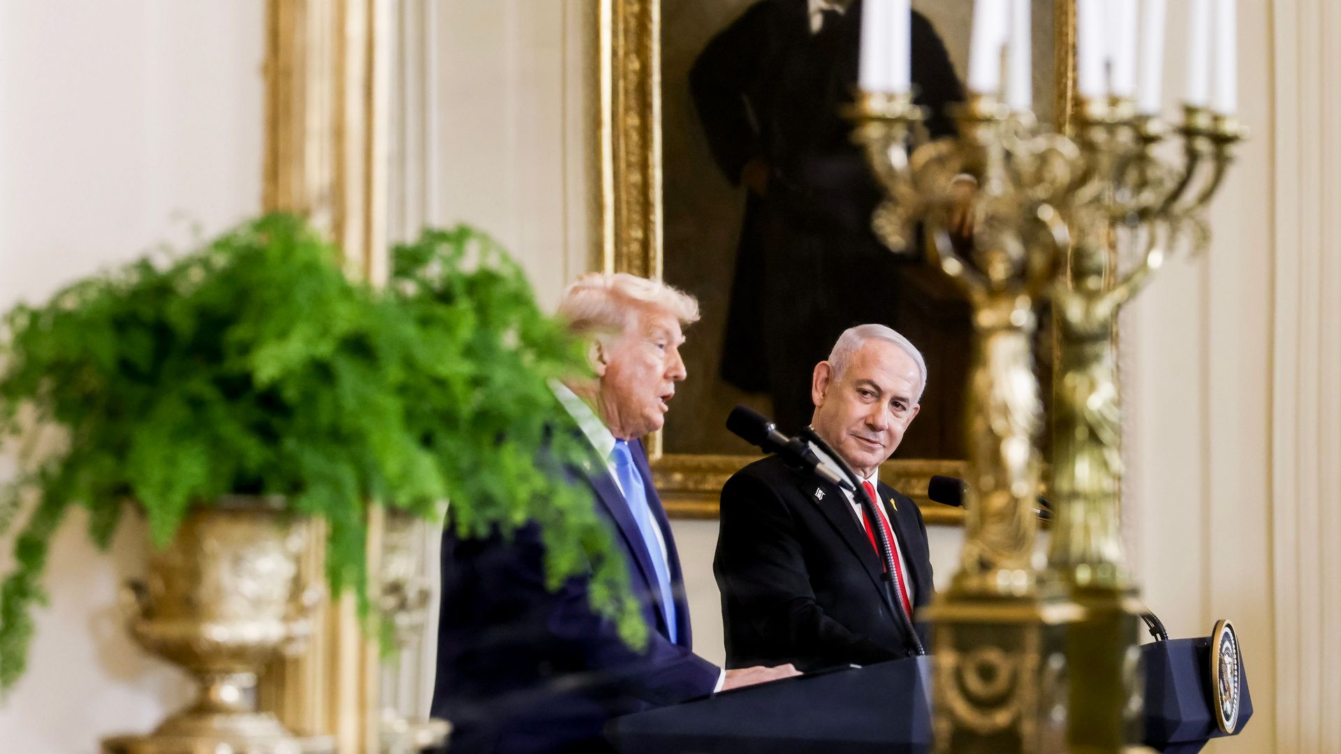 President Trump wearing a dark blue suit and Israeli Prime Minister Benjamin Netanyahu wearing a dark gray suit, standing at podiums in front of a large portrait in a white room with a gold candelabra and a plant in the foreground.