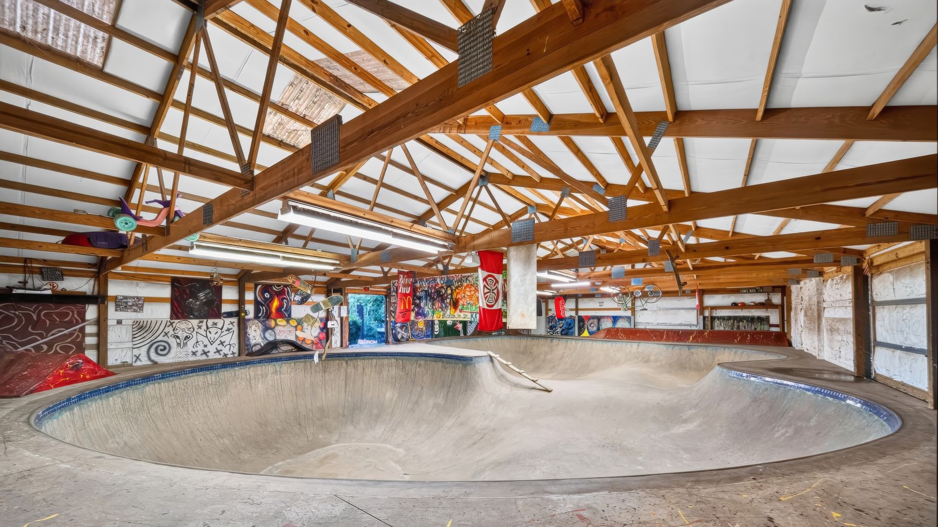 Indoor skatepark with a timber beam ceiling and white panels. A large curved concrete bowl dominates the foreground, with graffiti walls and hanging banners in the background.
