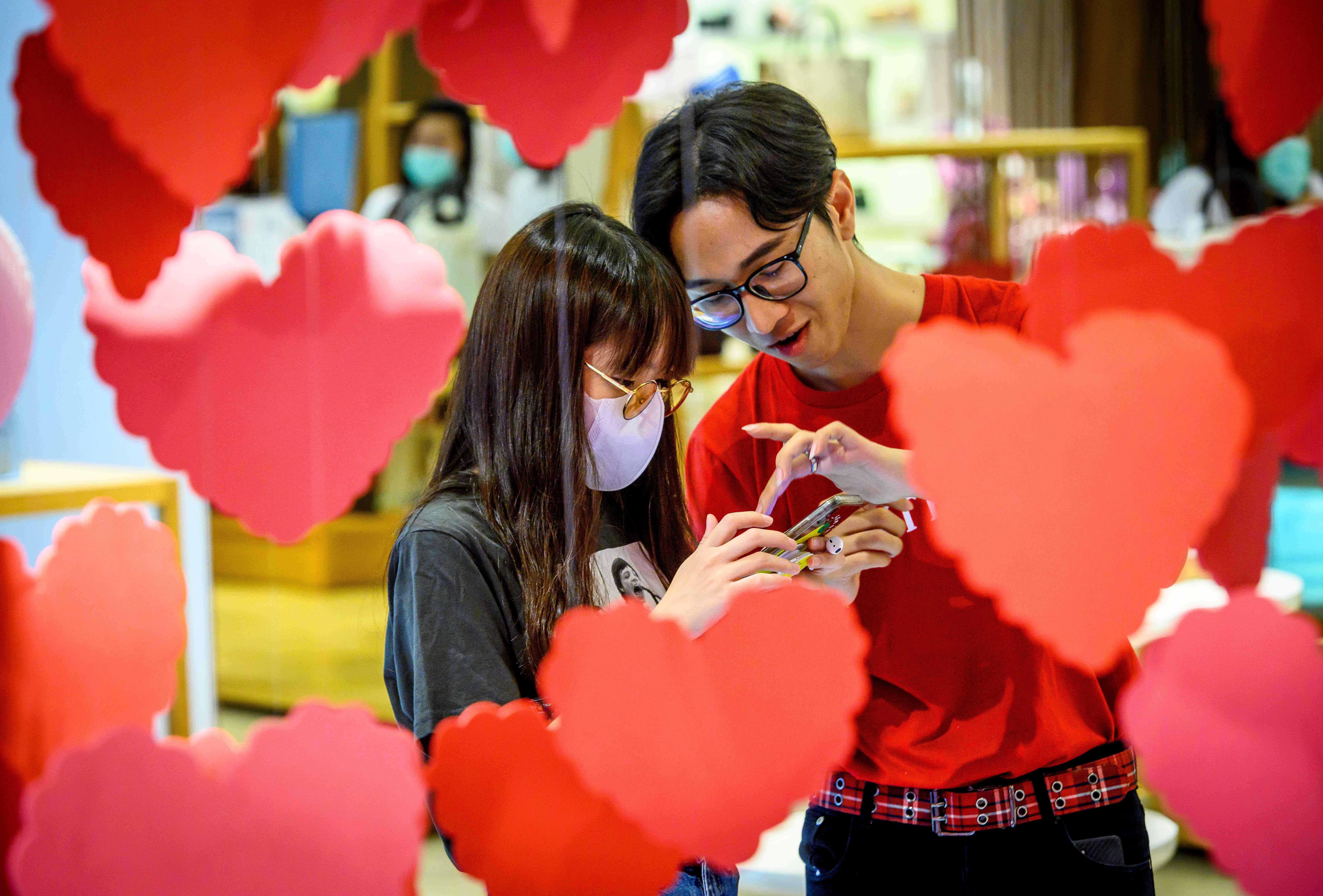 In this image, two people stand behind red cardboard hearts and one wears a face mask