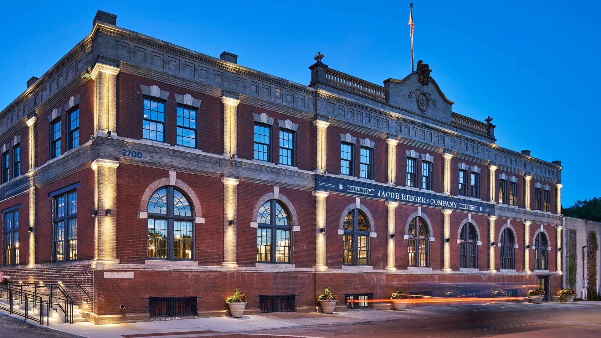 Evening exterior of a historic red brick building with arched windows and cream trim, warmly lit. A flag on top, 2700 on the side, and Jacob Rieger & Company signage across the facade.