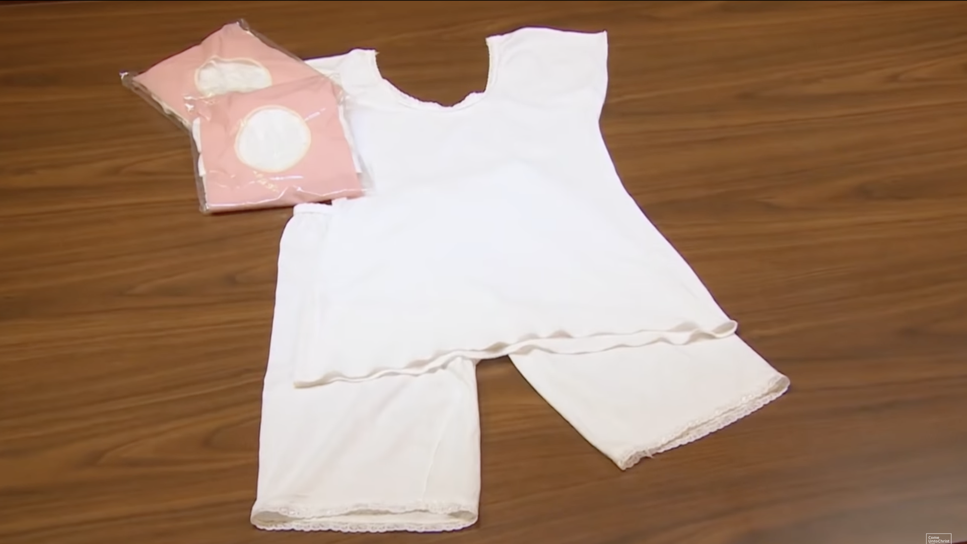 An image of Latter-day Saint temple garments: white shorts and a short-sleeved shirt, with two wrapped garment packages next to them on a wood surface.