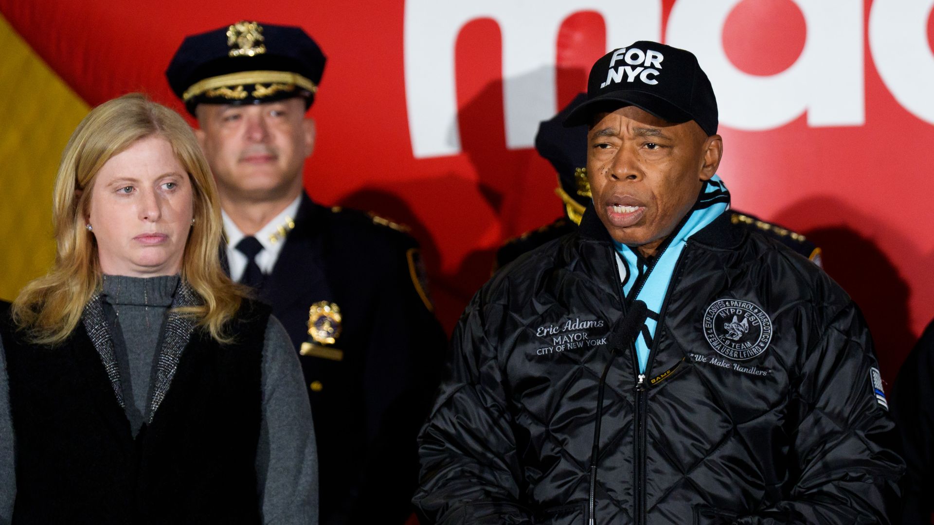 A fair-skinned NYPD Commissioner Jessica Tisch, wearing long, strawberry blonde hair, a gray sweater and black coat, and NYC Mayor Eric Adams, wearing a black cap with the words "FOR NYC" on it, a black coat and blue scarf, at a podium during a briefing.