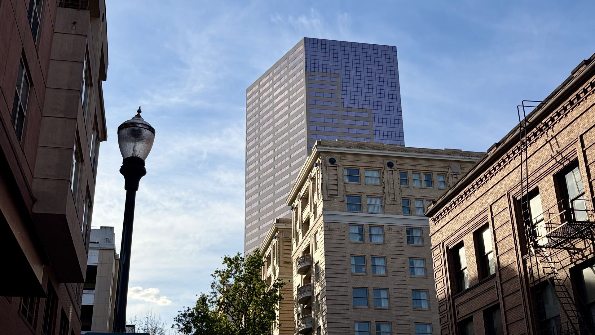 City street scene shows a tall pink-glass office building between brown brick facades. A vintage streetlamp stands in the foreground left, with fire escapes along the right and a blue sky above.