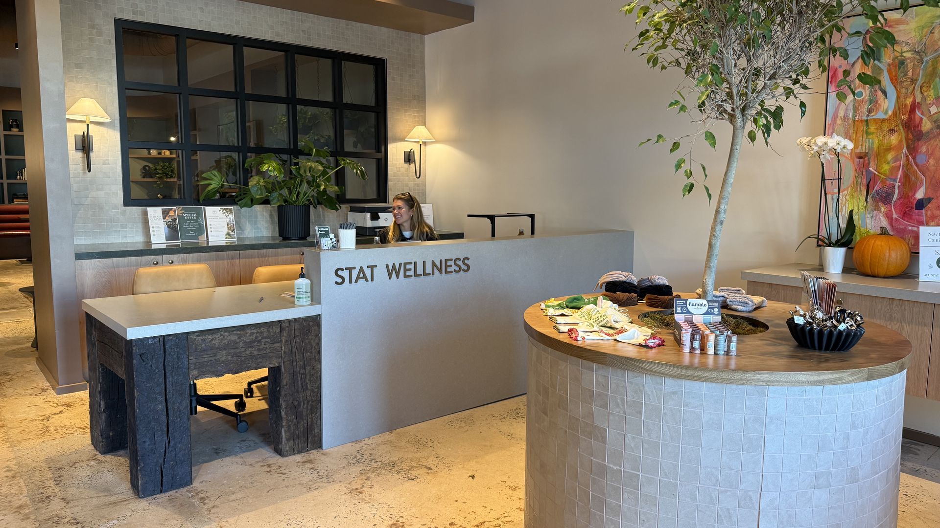 Reception at STAT WELLNESS: curved tiled desk, a smiling receptionist behind a glass partition, with potted plants, a tall indoor tree, and colorful wall art near a round product display.