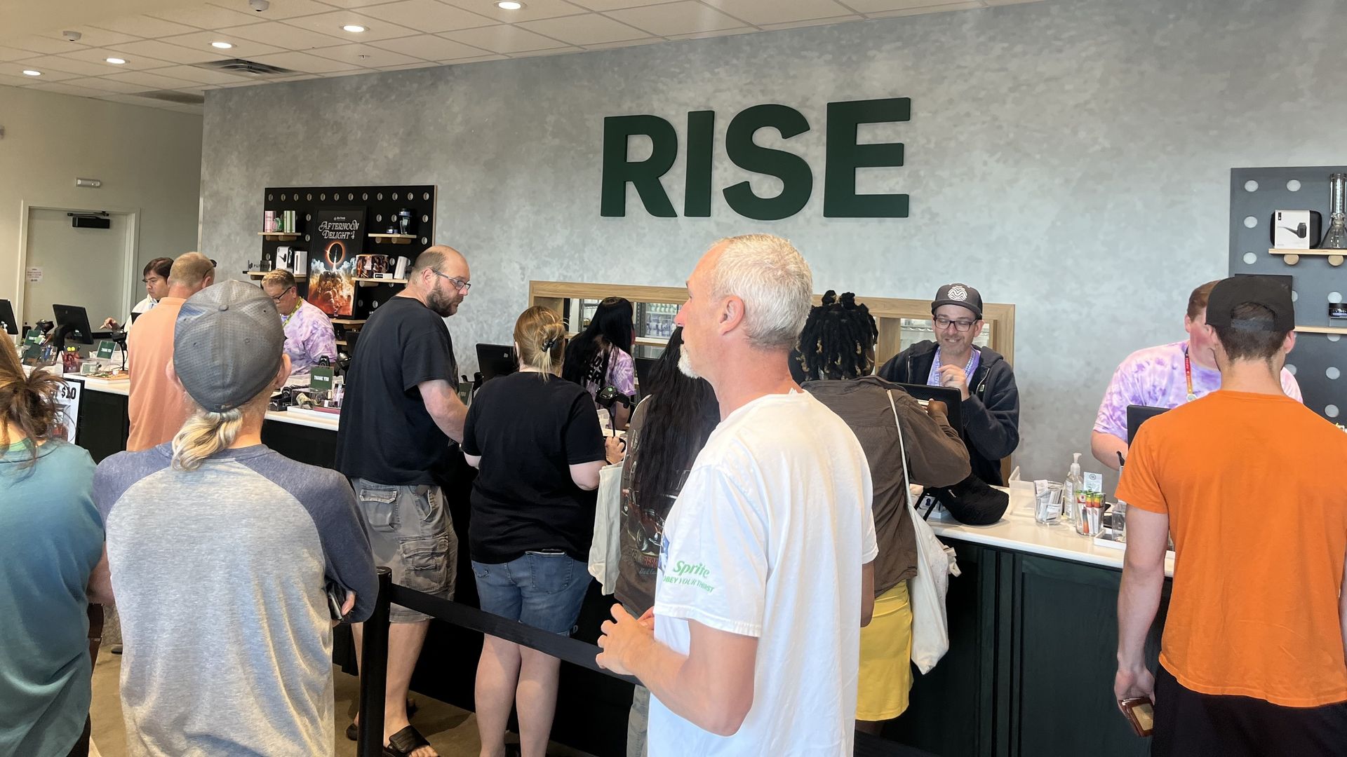 Interior of a busy shop or cafe with people standing in line at the counter. Large green letters spelling RISE on a gray textured wall behind the counter.