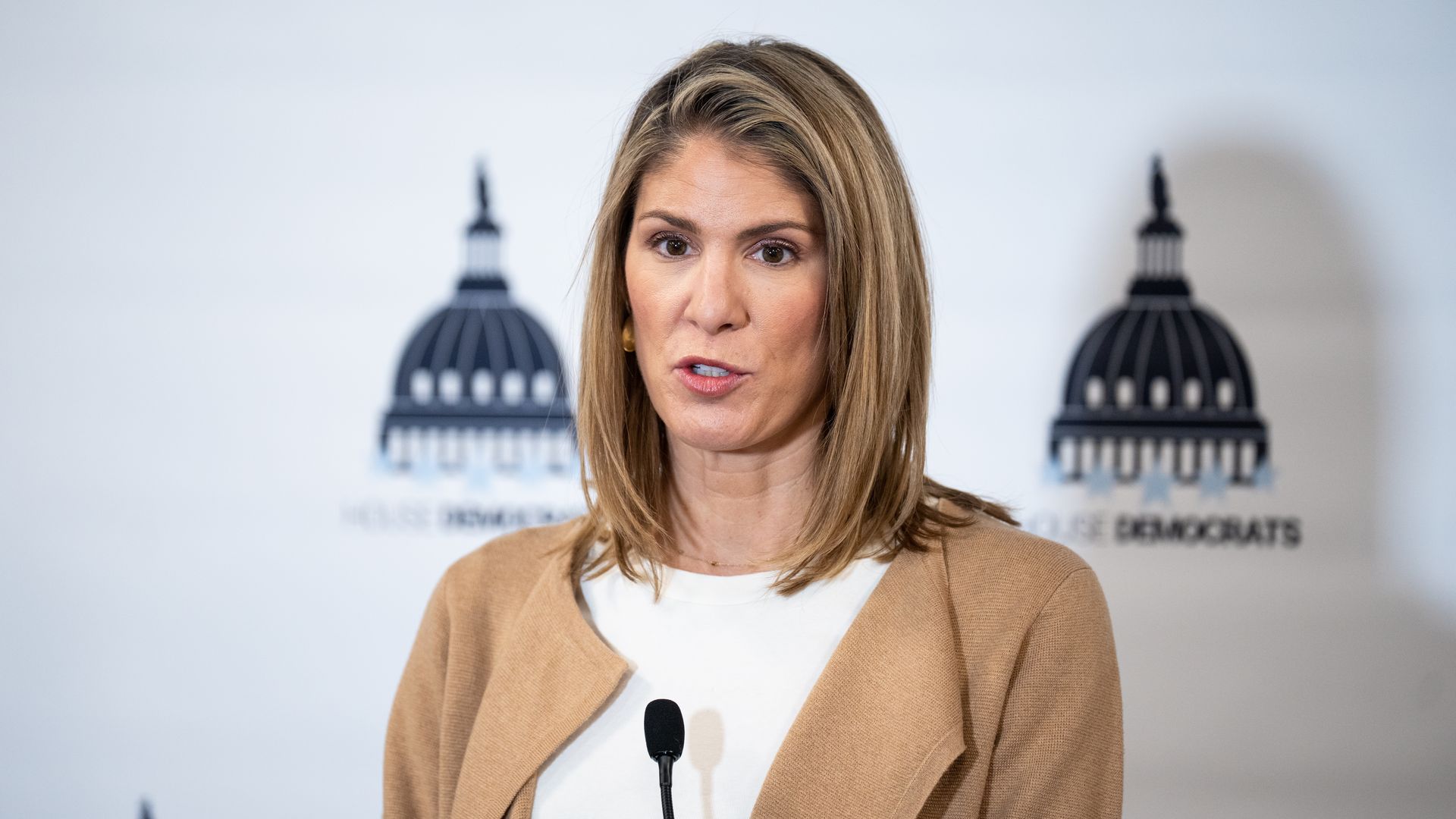 Rep. Lori Trahan, wearing a beige pullover over a white dress, speaks at a microphone in front of a "House Democrats backdrop.