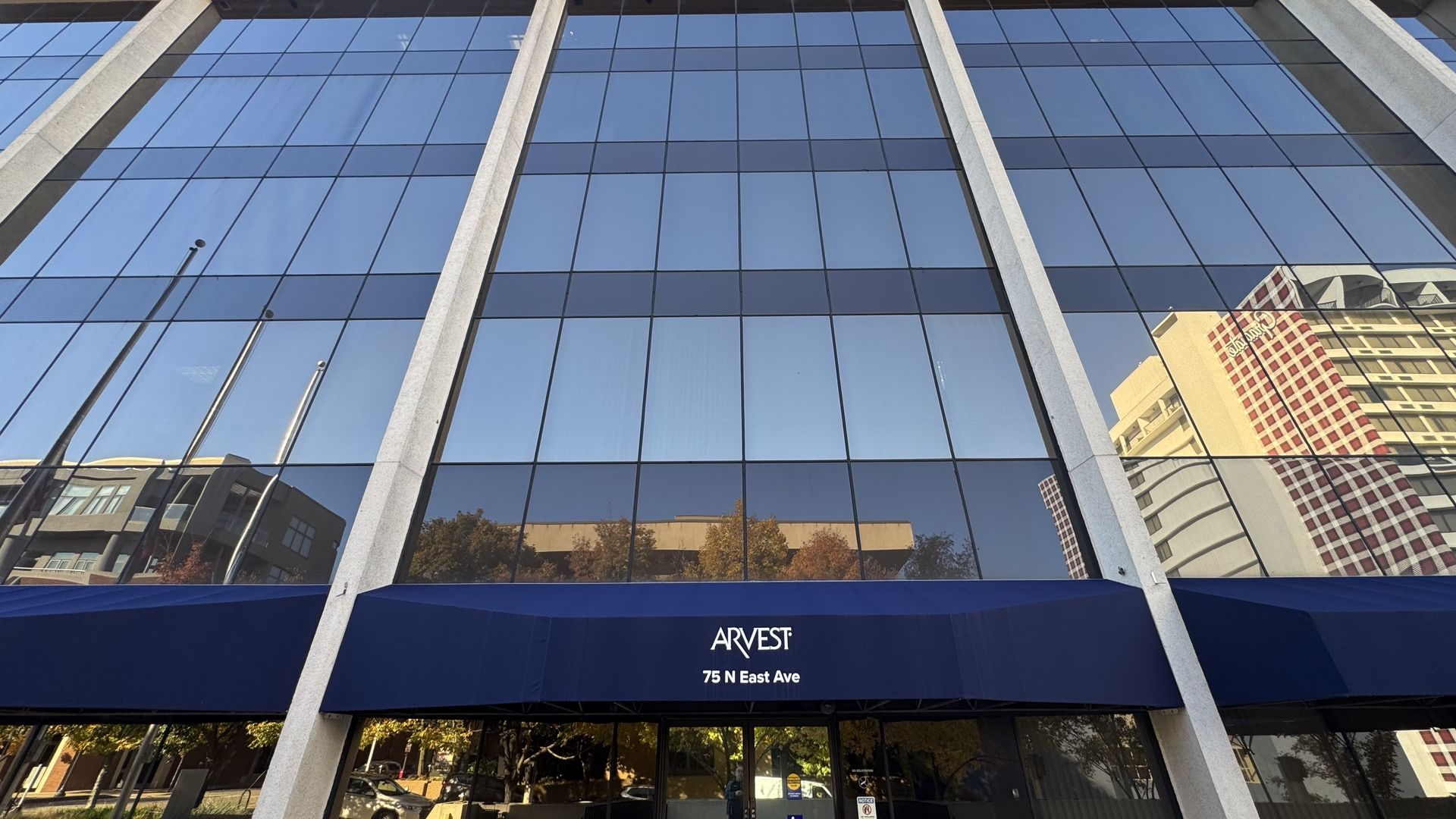 Tall modern office building with large reflective blue-tinted windows, white pillars, and a blue awning reading "ARVEST 75 N East Ave" against a clear sky and city reflection.