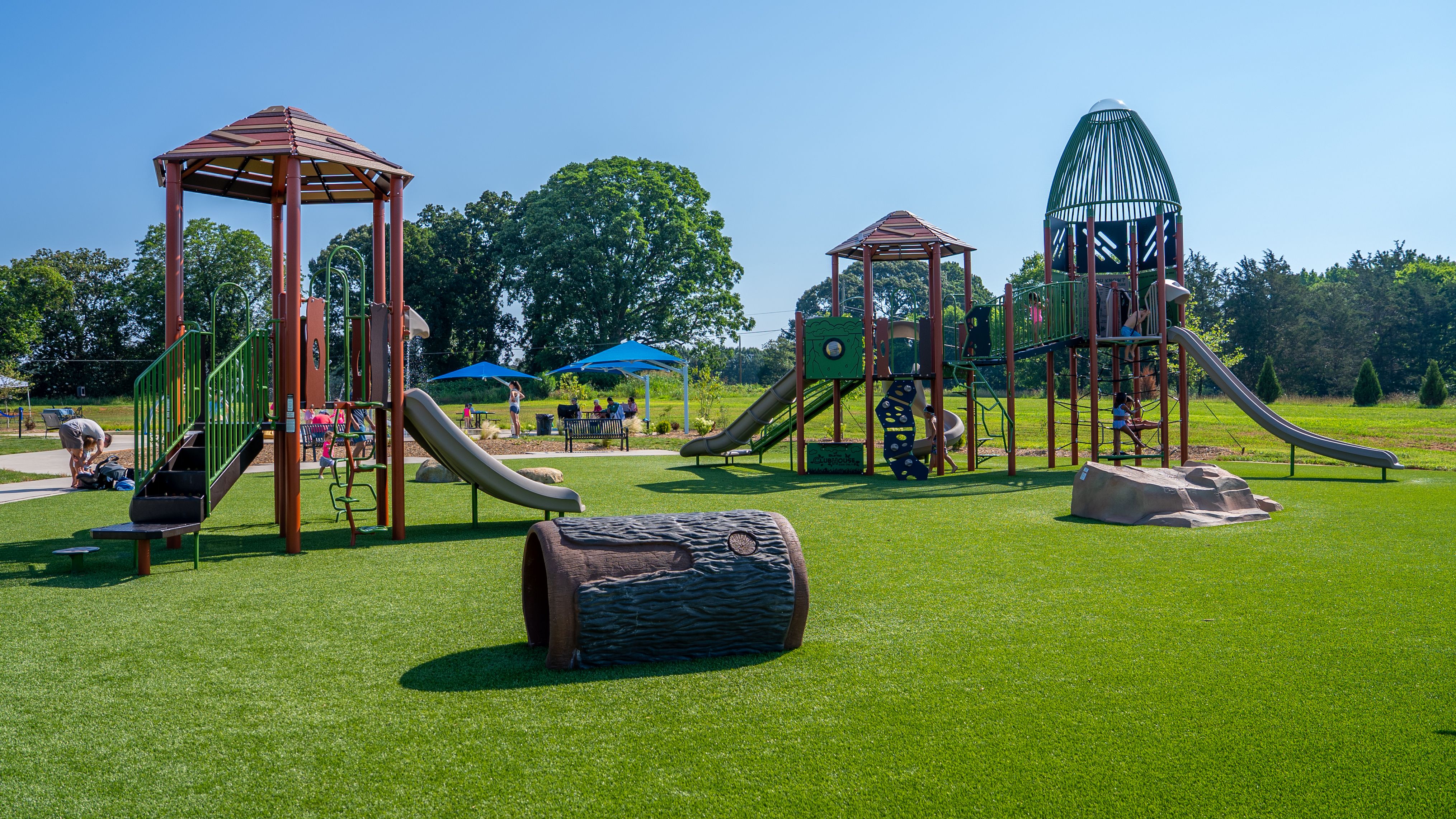 Sunny park playground with red-brown structures, green slides, and a tall dome climber. Children play on equipment while blue shade umbrellas and benches sit beside a grassy field with trees.