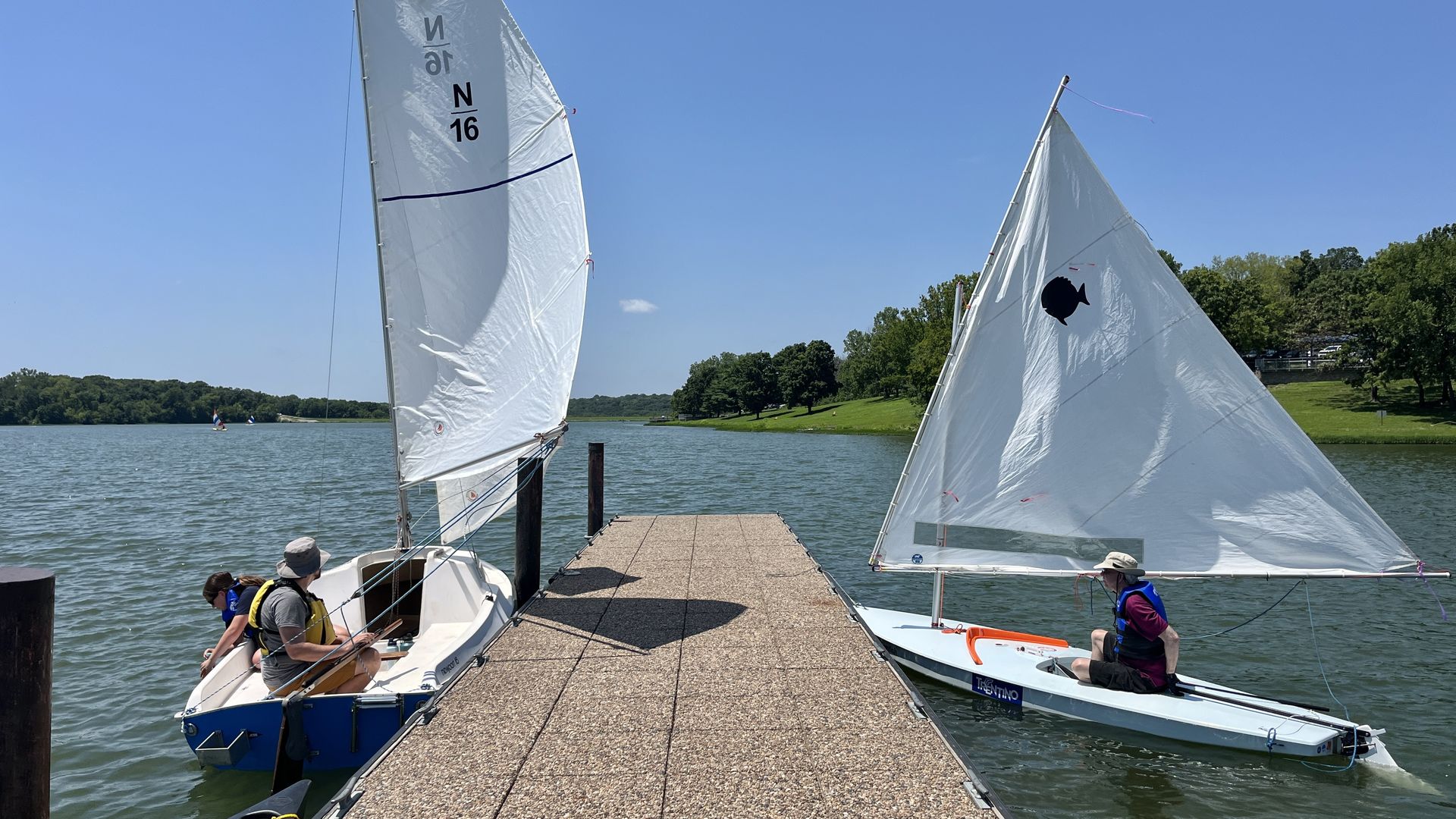 Two small sailboats docked beside a pier on a lake with clear blue sky and green trees in the background. One boat holds two people, the other one person, all wearing life jackets.