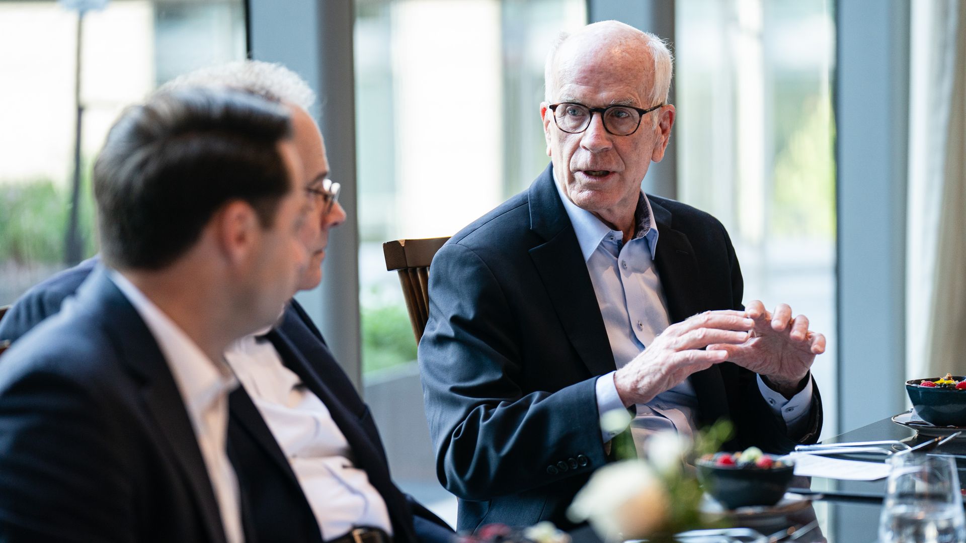 Two men in suits chat at a sunlit cafe table; the older man with glasses gestures while the other man sits in the foreground, with plates and bowls visible on the table.