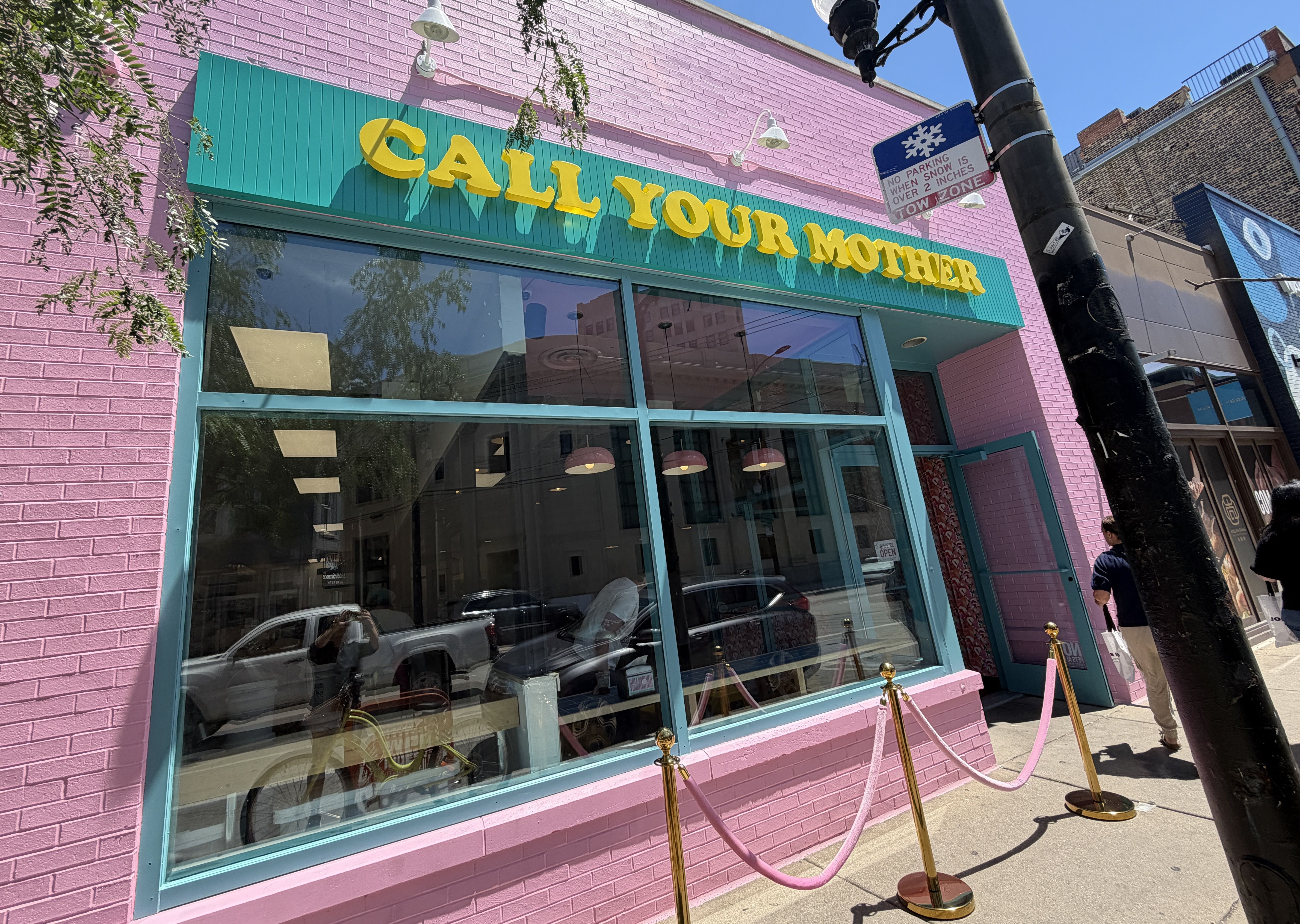 A pink brick storefront with a turquoise awning reading CALL YOUR MOTHER in large yellow letters. Glass windows reflect the street; a lamppost and a no-parking sign are nearby.