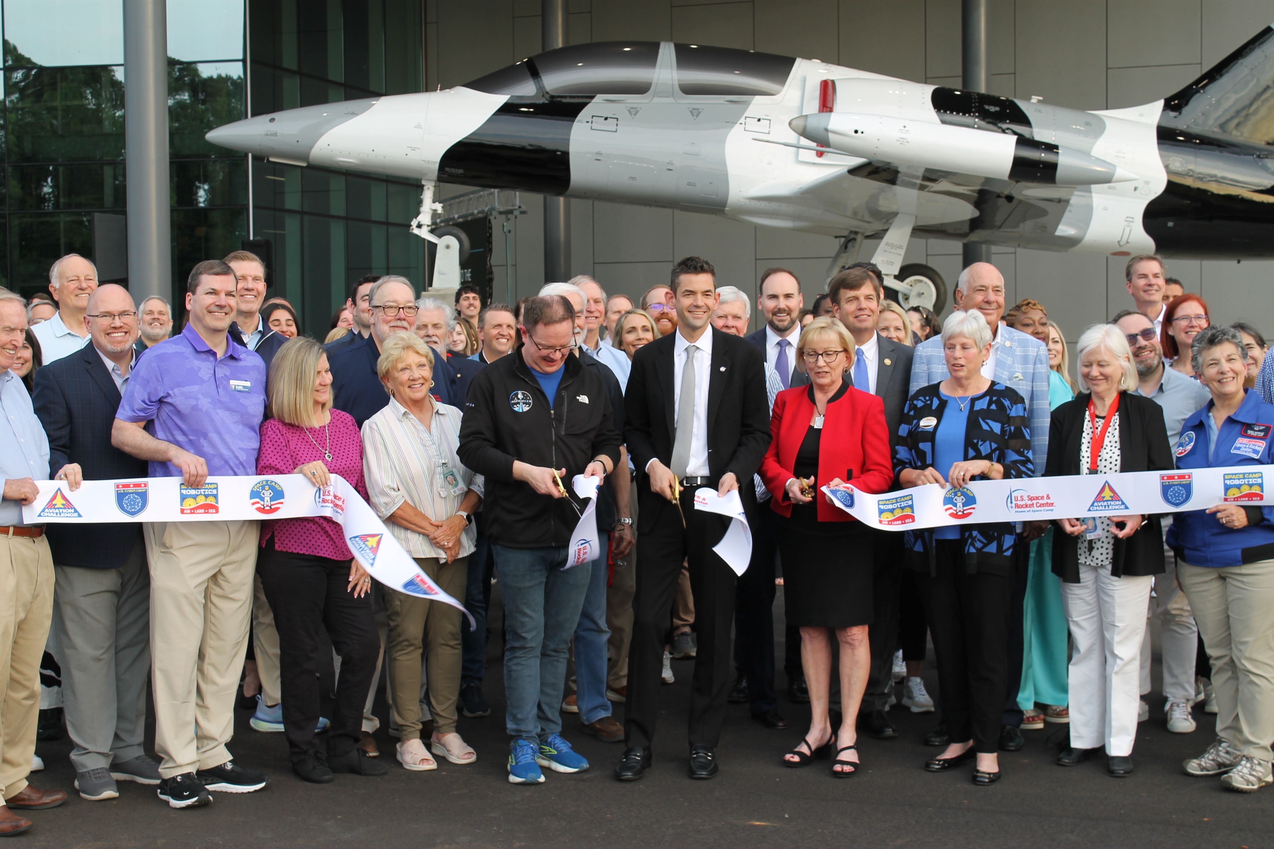Diverse group of people posing for a ribbon-cutting photo in front of a large, futuristic white and gray aircraft on display outside a modern building.