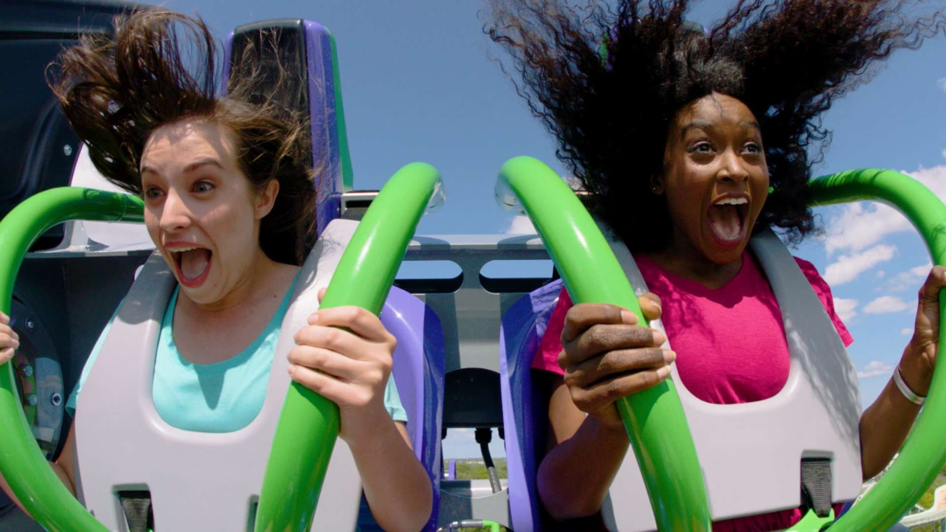 Two women on a roller coaster scream, gripping bright green safety bars. One wears a teal top, the other a pink shirt, with a clear blue sky behind them.