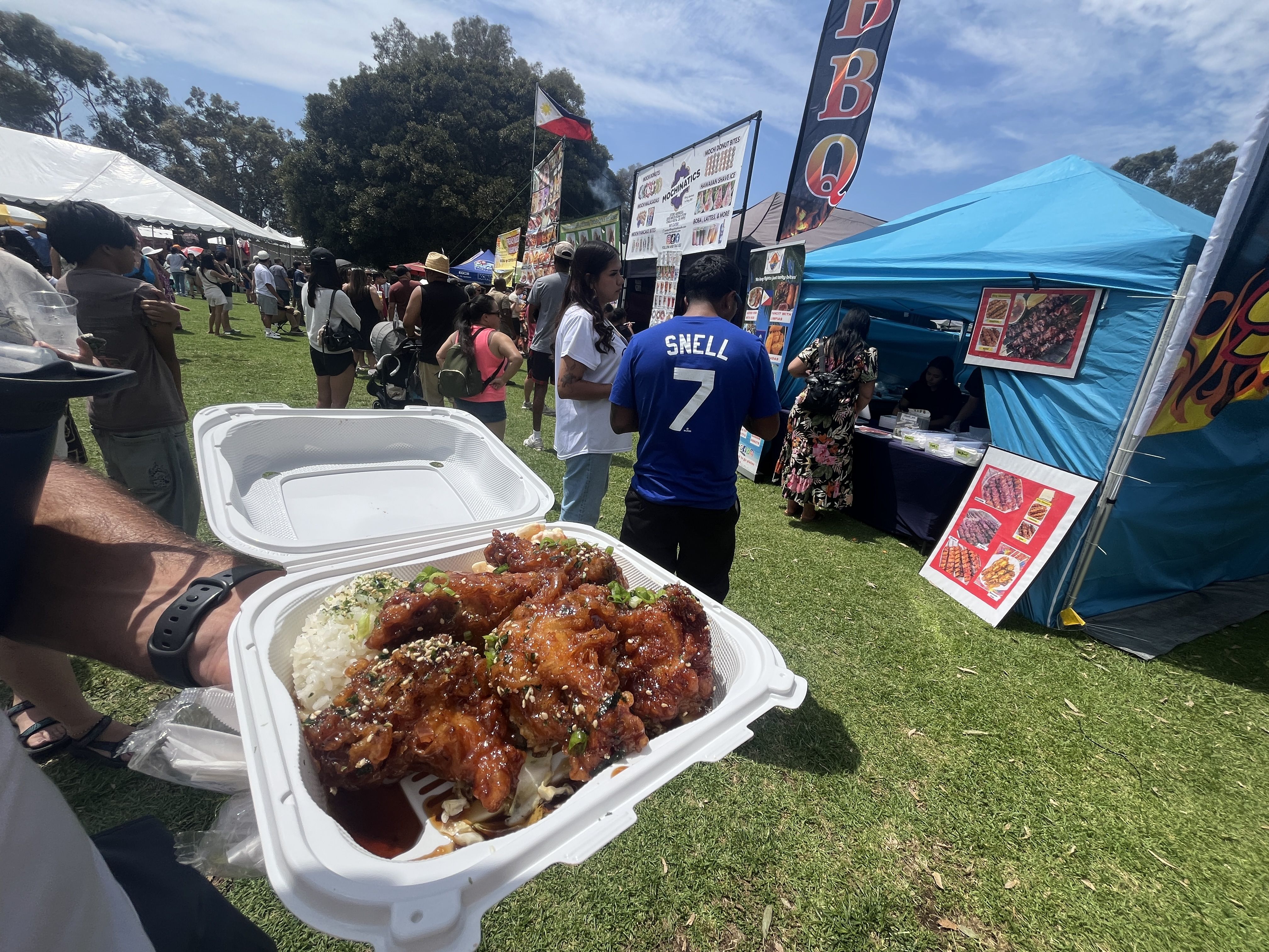 Outdoor food festival with people in line near booths under blue tents. Foreground shows a hand holding a white container with glazed chicken wings, rice, and garnish.