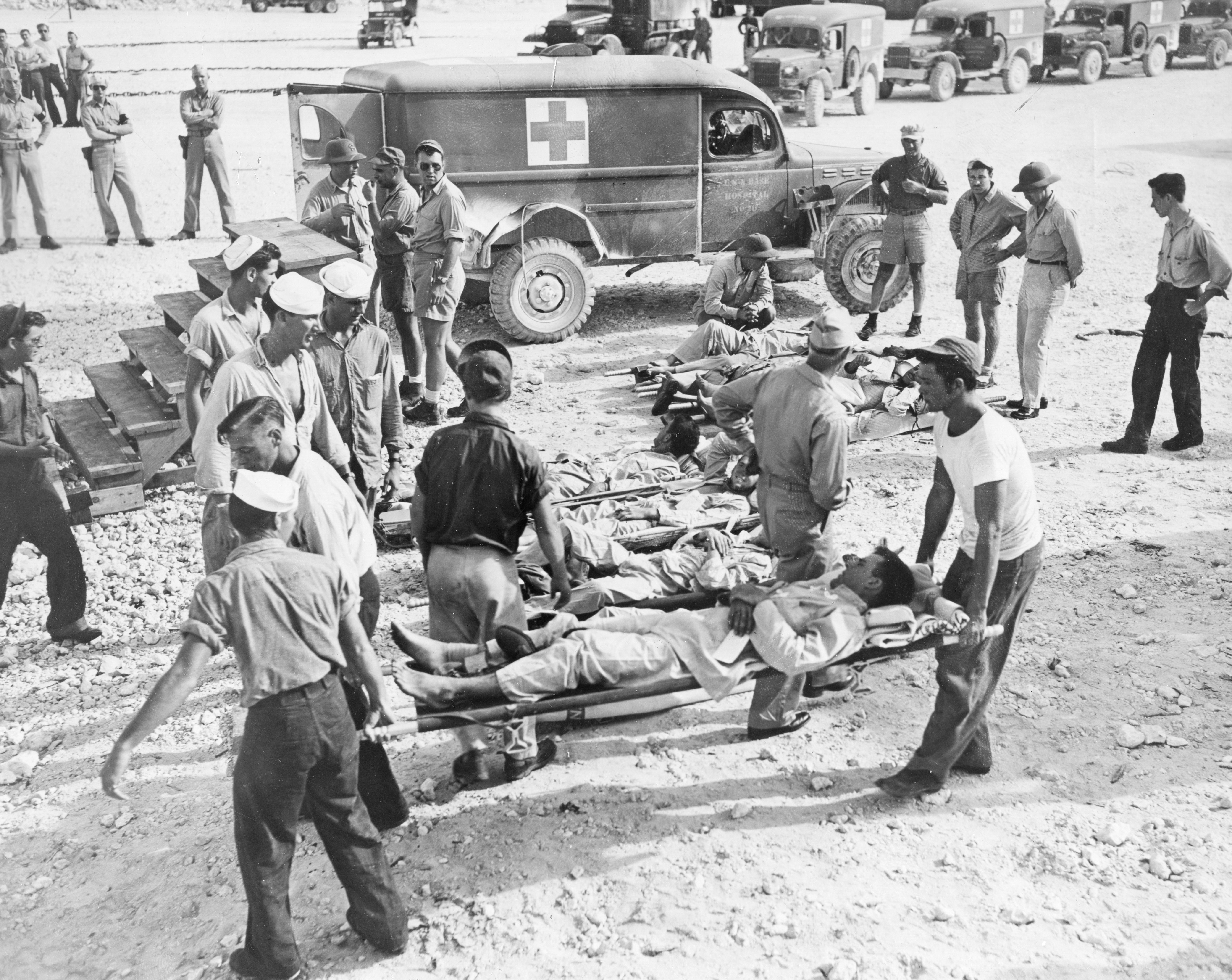 Black-and-white photo of soldiers carrying wounded comrades on stretchers near military ambulances, with others standing or observing on rocky ground in a wartime setting.