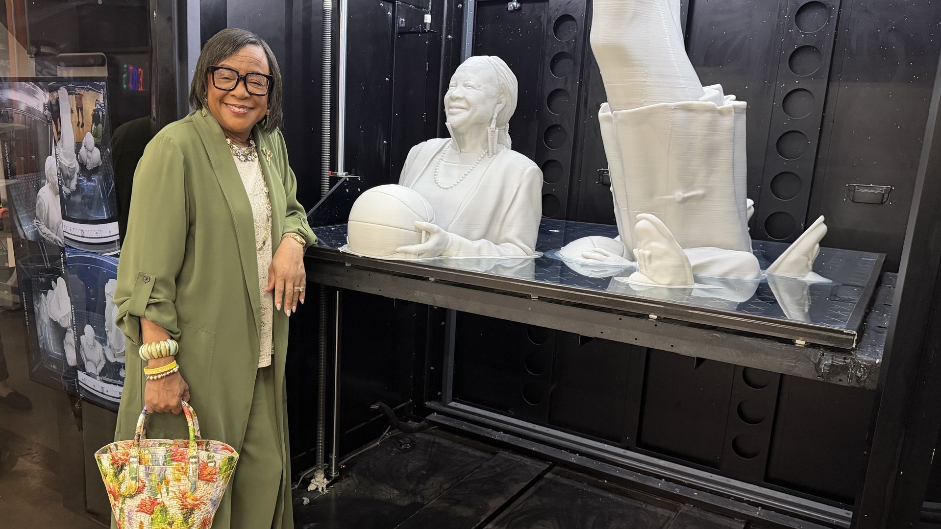A smiling woman in a green blazer with a colorful tote stands beside a life-size white sculpture of a person in a blazer wearing pearls, holding a basketball, inside a large black 3D‑printing enclosure.