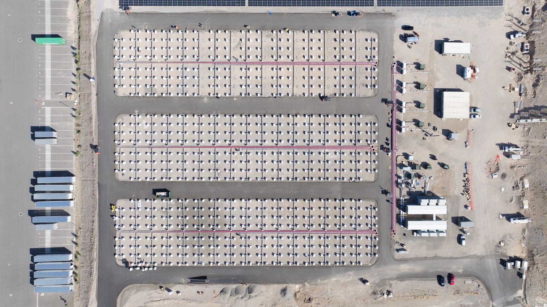 Aerial view of a large battery farm on a gray paved surface, adjacent to a parking area with trucks and equipment and next to a solar panel system