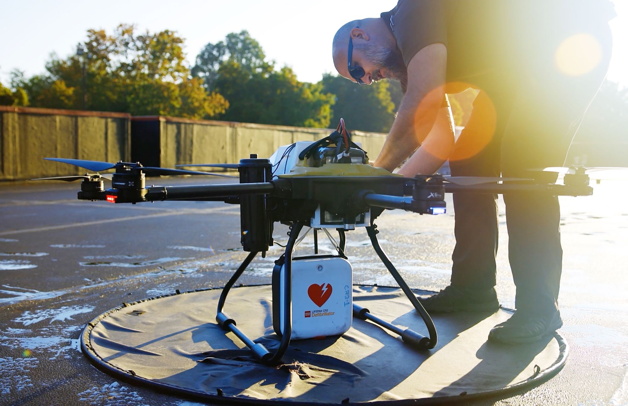 A man wearing sunglasses sets up a large black drone equipped with a white defibrillator case on an outdoor landing pad with trees and sunlight in the background.