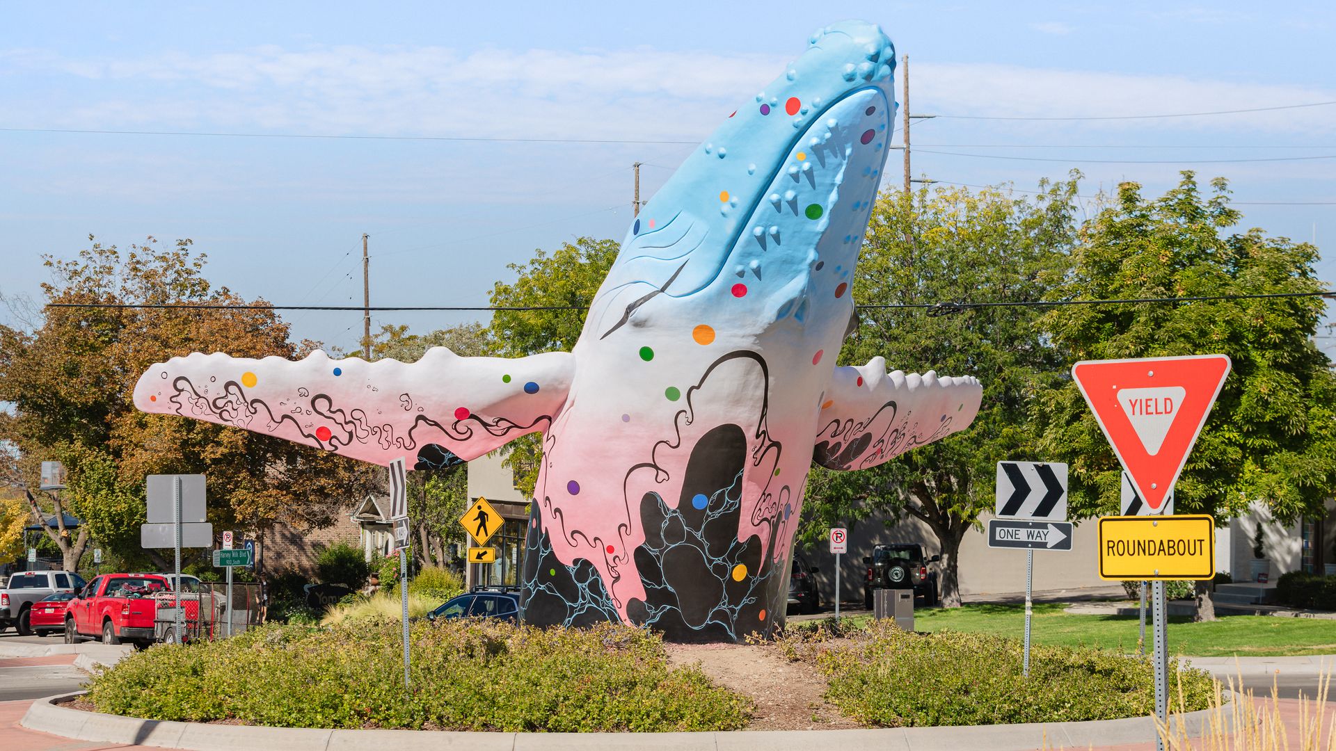 Large, colorful sculpture of a sea creature rising from a roundabout, painted light blue with multicolor dots and dark outlines, surrounded by trees, cars, and a yield sign under a blue sky.