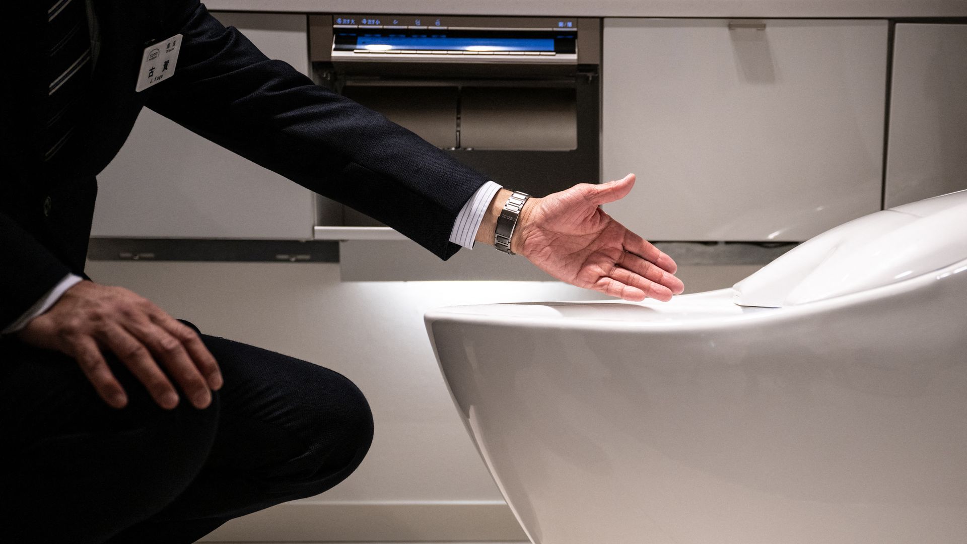 Person in a dark suit reaches toward a white toilet in a modern bathroom; silver wristwatch visible.
