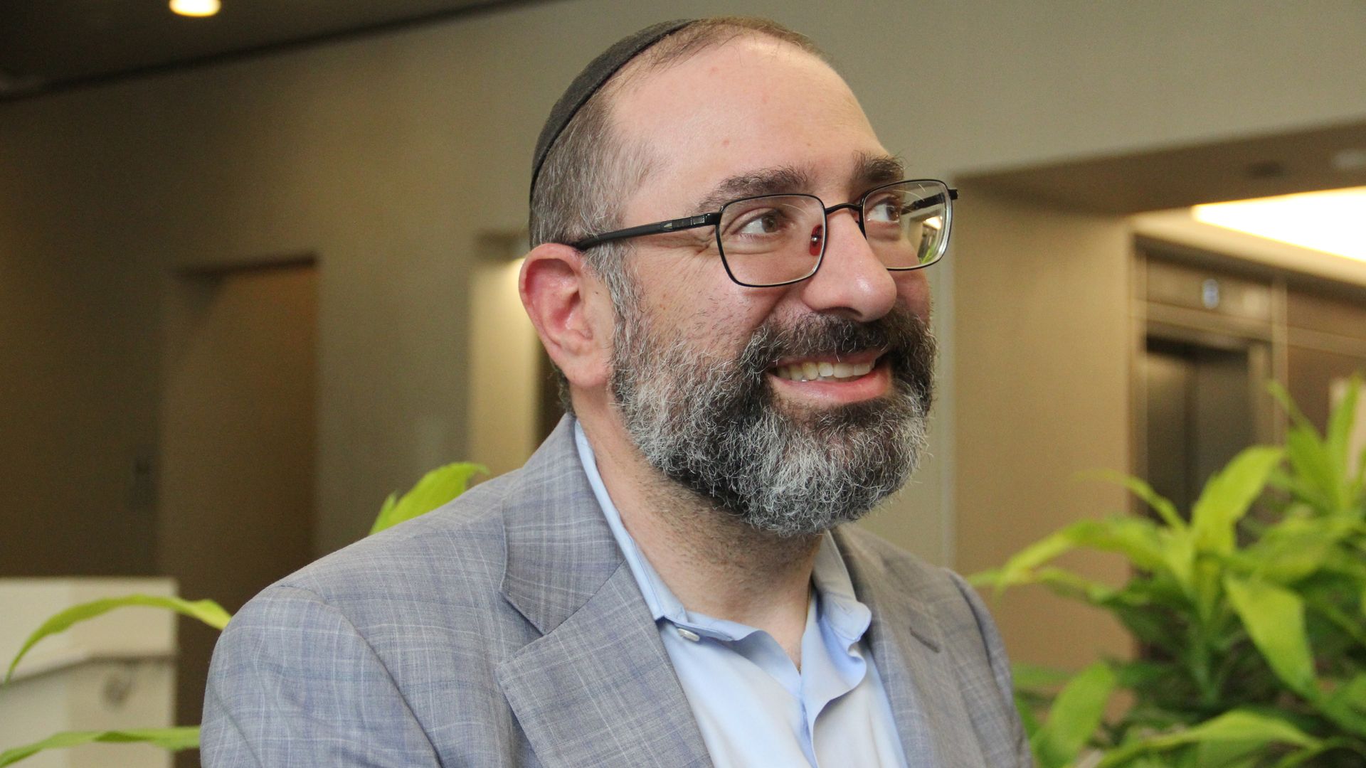 Portrait of a smiling man with a gray beard and glasses, wearing a black kippah and a gray plaid suit, indoors with plants in the background.