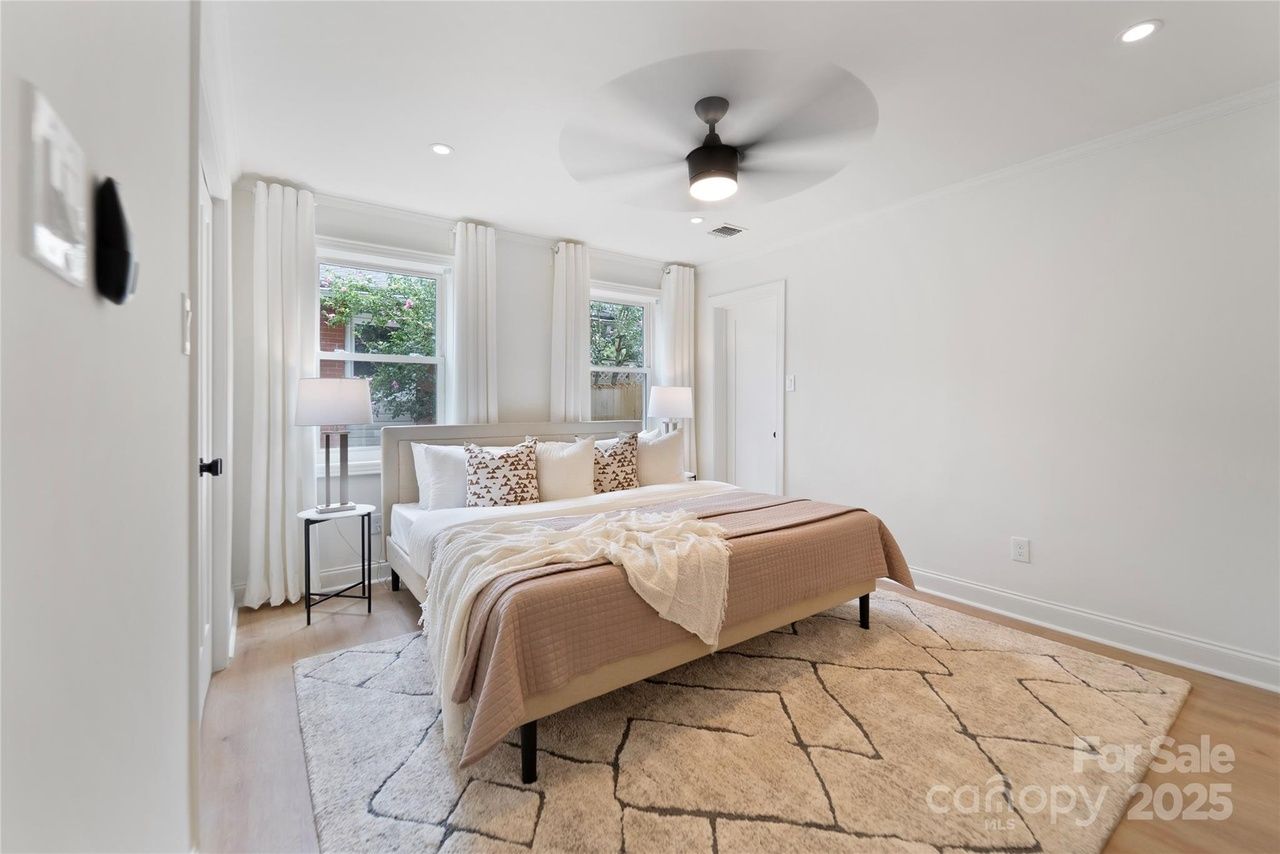 Bright bedroom with white walls, two windows with white curtains, a beige bed with patterned and white pillows, a beige quilt, two bedside lamps, a ceiling fan, and a geometric patterned rug.