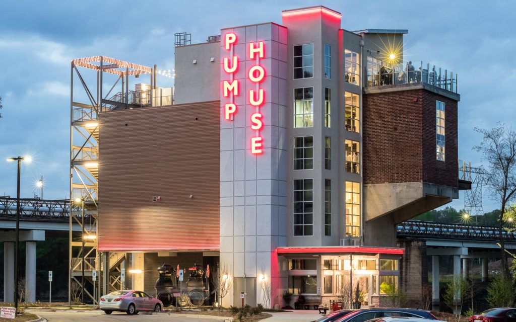 The Pump House, a modern multi-story building with red neon sign, large windows, rooftop deck, and a parking lot with cars, under a cloudy evening sky.