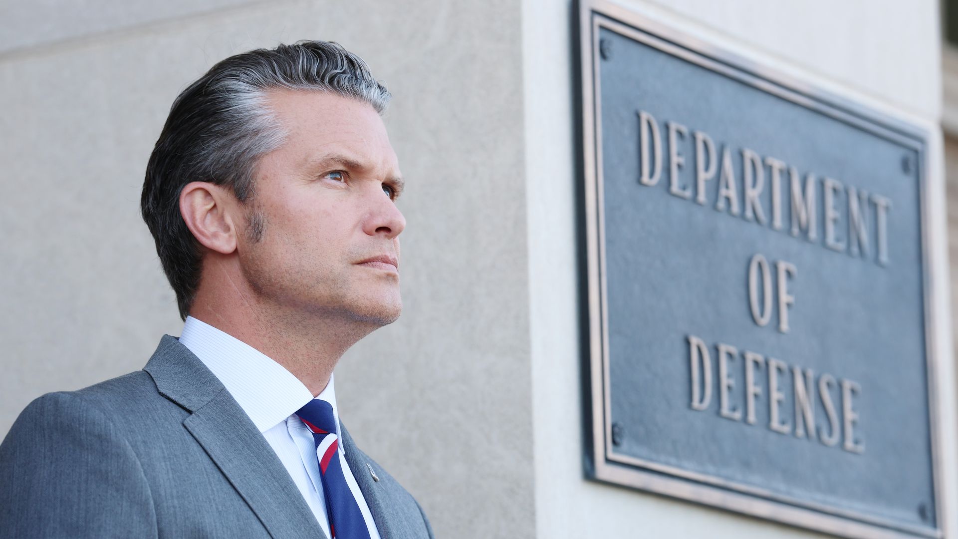 Pete Hegseth stares forward in a gray suit next to a sign that reads "DEPARTMENT OF DEFENSE."
