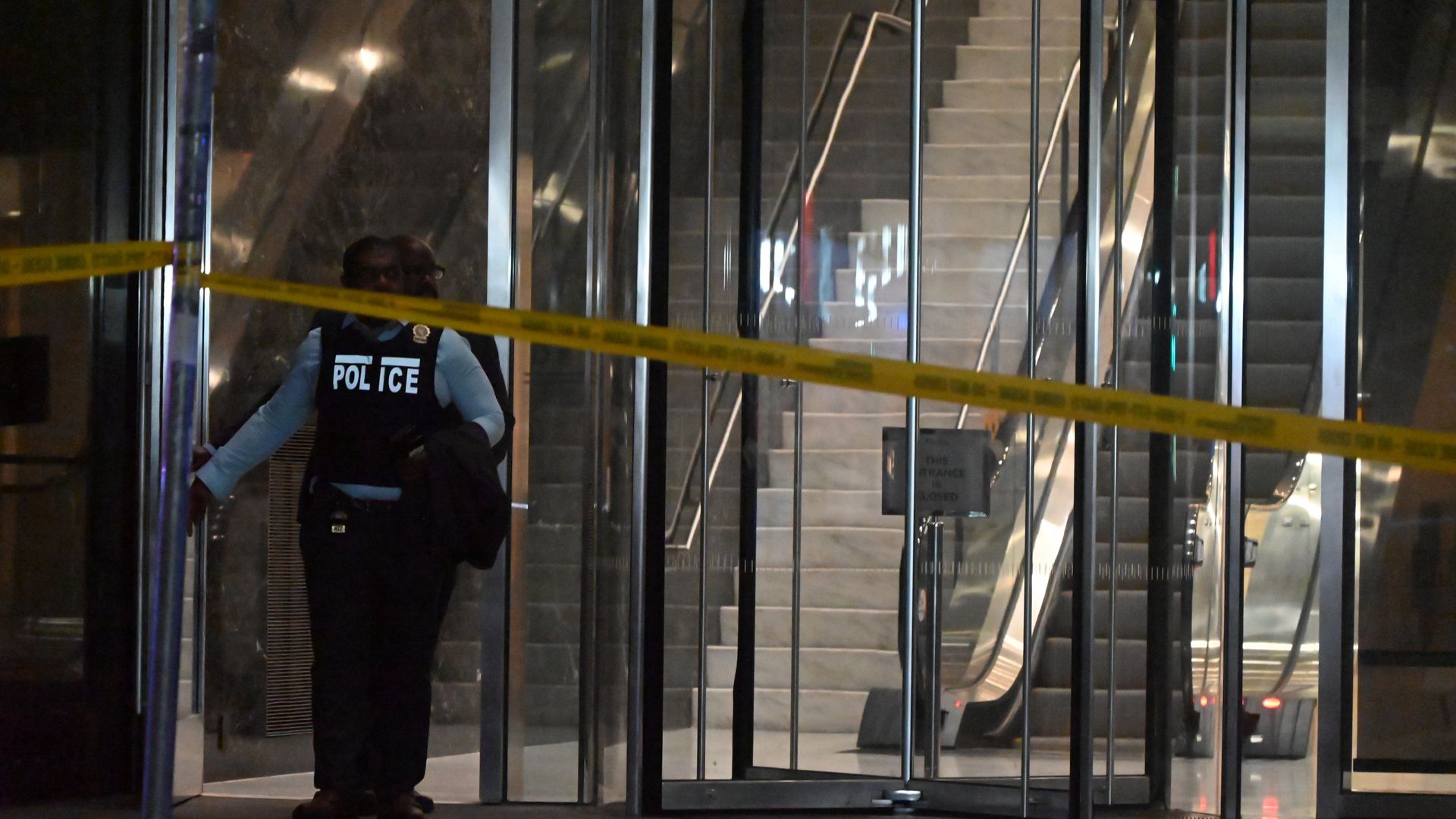 Police officer standing inside glass entrance of a building with yellow caution tape across, stairs and escalators visible inside, dimly lit scene.
