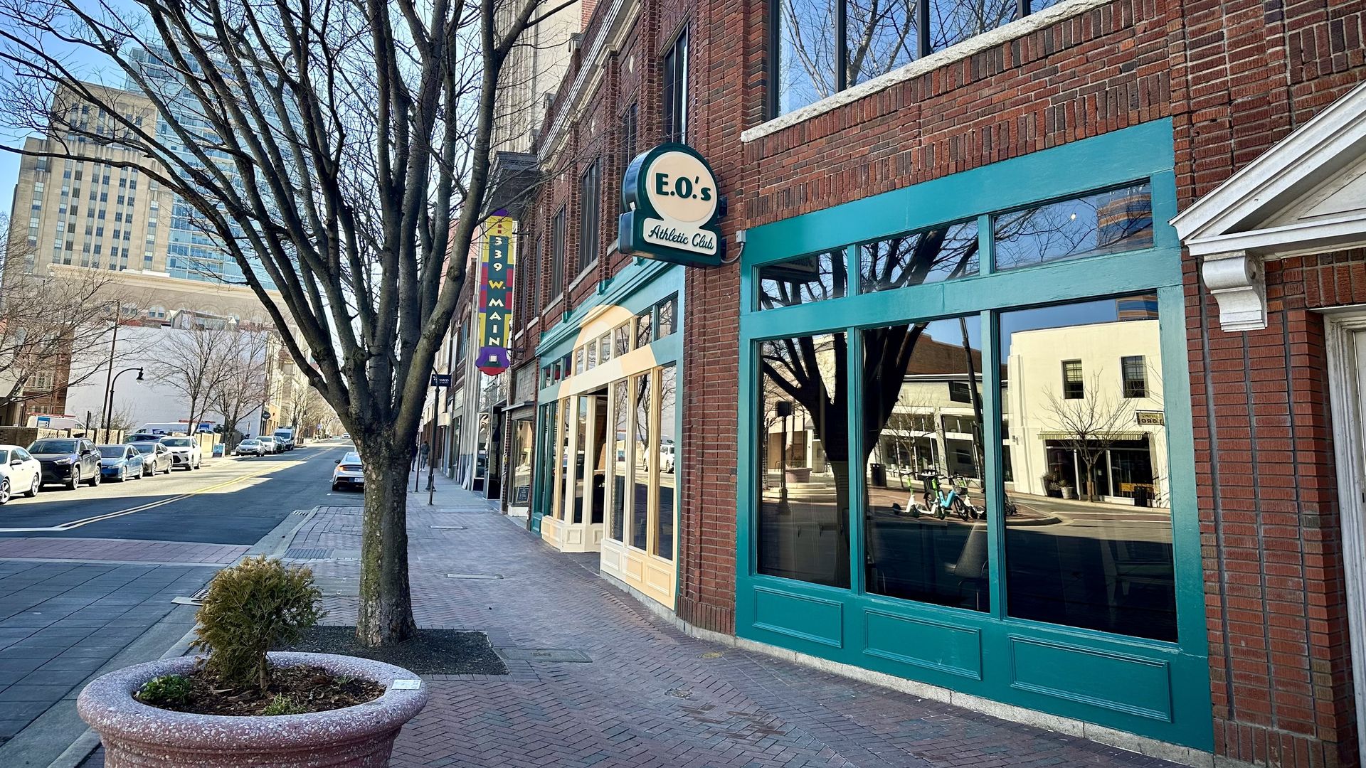 Brick building with teal-trimmed windows, sign reading "E.O.'s Athletic Club," leafless trees, blue sky, and reflections on glass along a quiet city sidewalk.