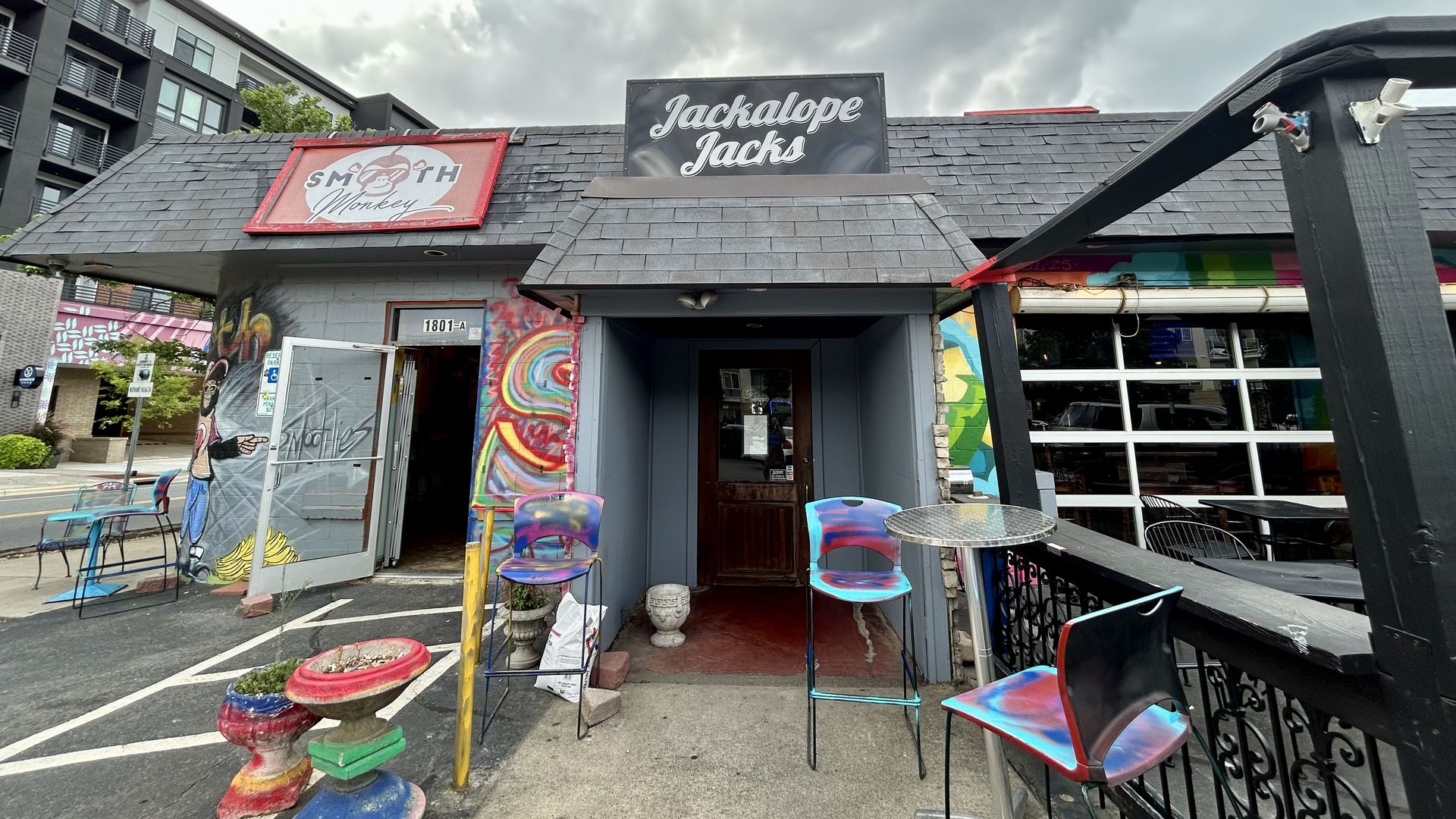 Exterior of Jackalope Jacks bar with colorful chairs and painted planters outside under a cloudy sky. Adjacent building has a Smooth Monkey sign with graffiti art.