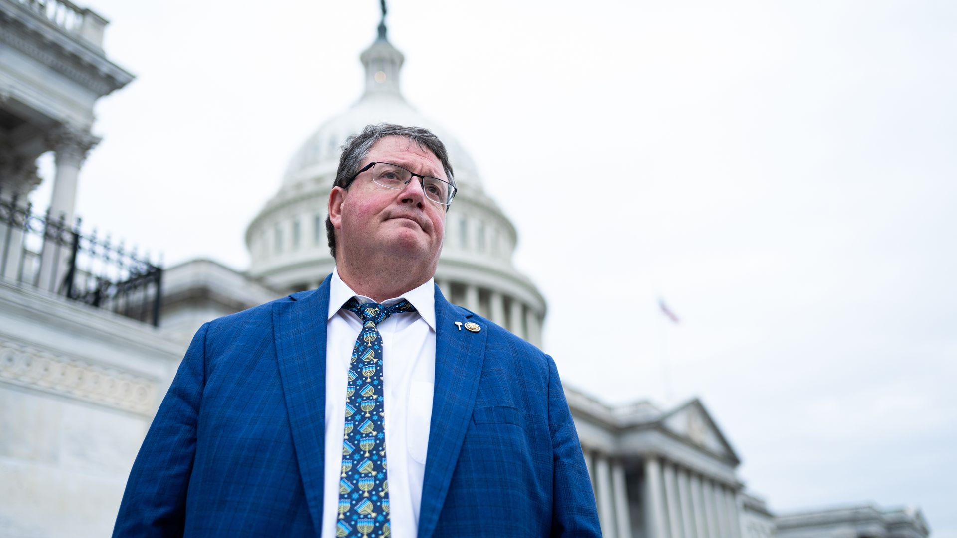 Man in a blue suit and Hanukkah-themed tie standing outside a white domed government building, looking slightly upward with a thoughtful expression on a cloudy day.