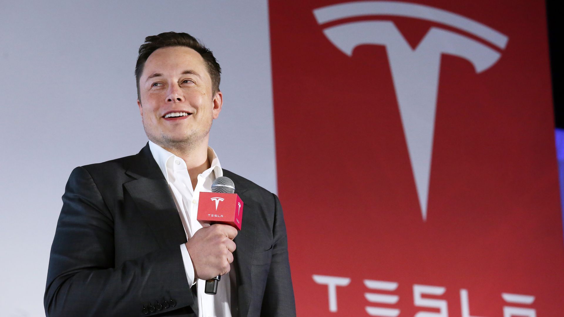In this image, Elon Musk stands in a suit with a microphone in front of a red sign depicting the Tesla logo.