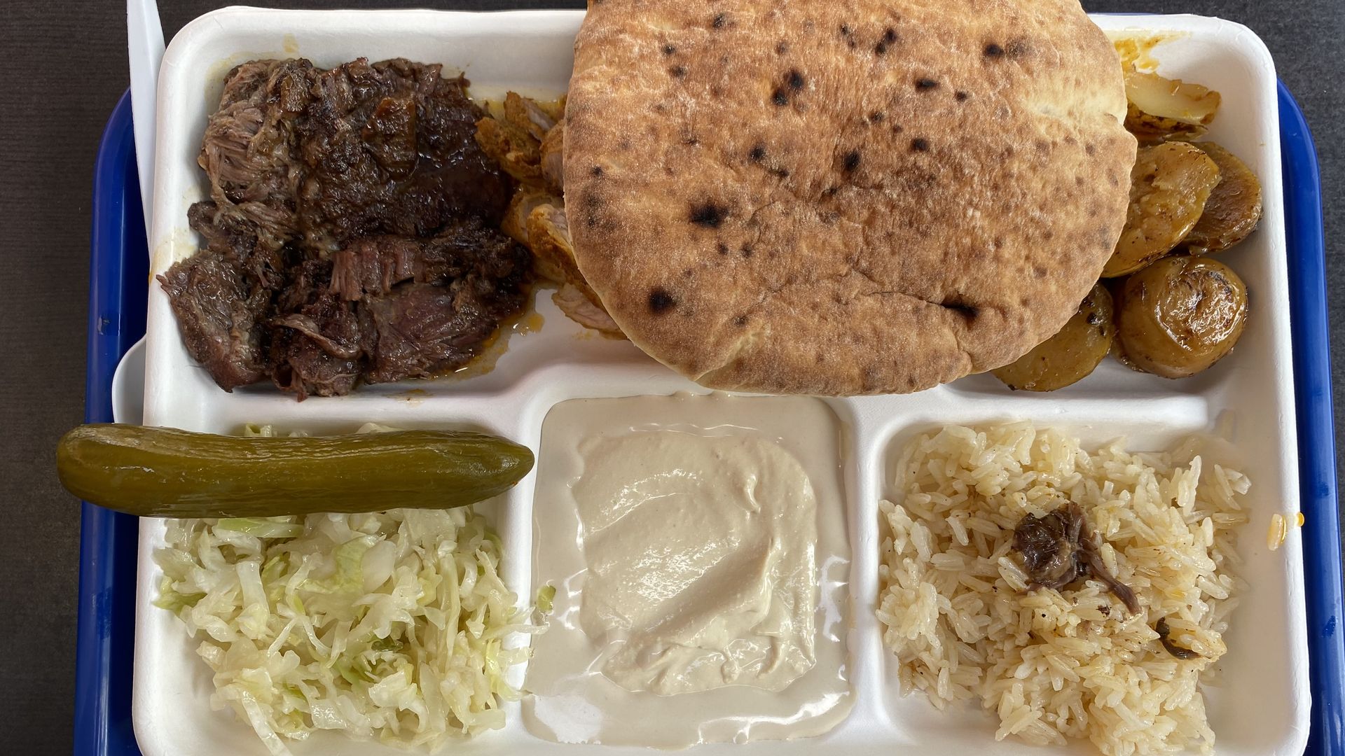 A compartmentalized tray filled with beef, chicken, potatoes, rice, shredded cabbage, tahini, a pickle and a pita