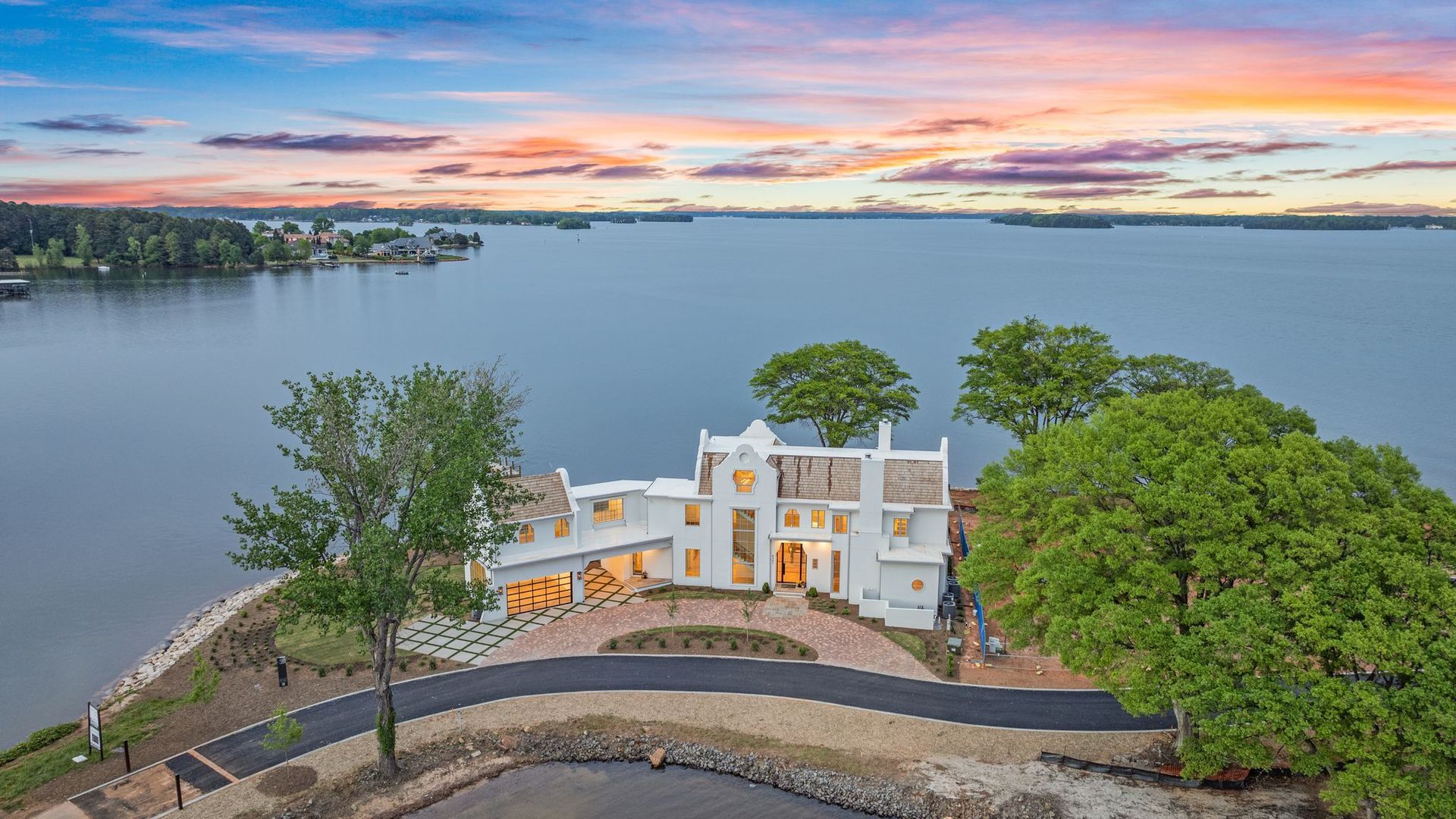 White two-story waterfront house with large windows and a garage, surrounded by green trees, set against a calm lake and a colorful sunset sky with pink and orange hues.