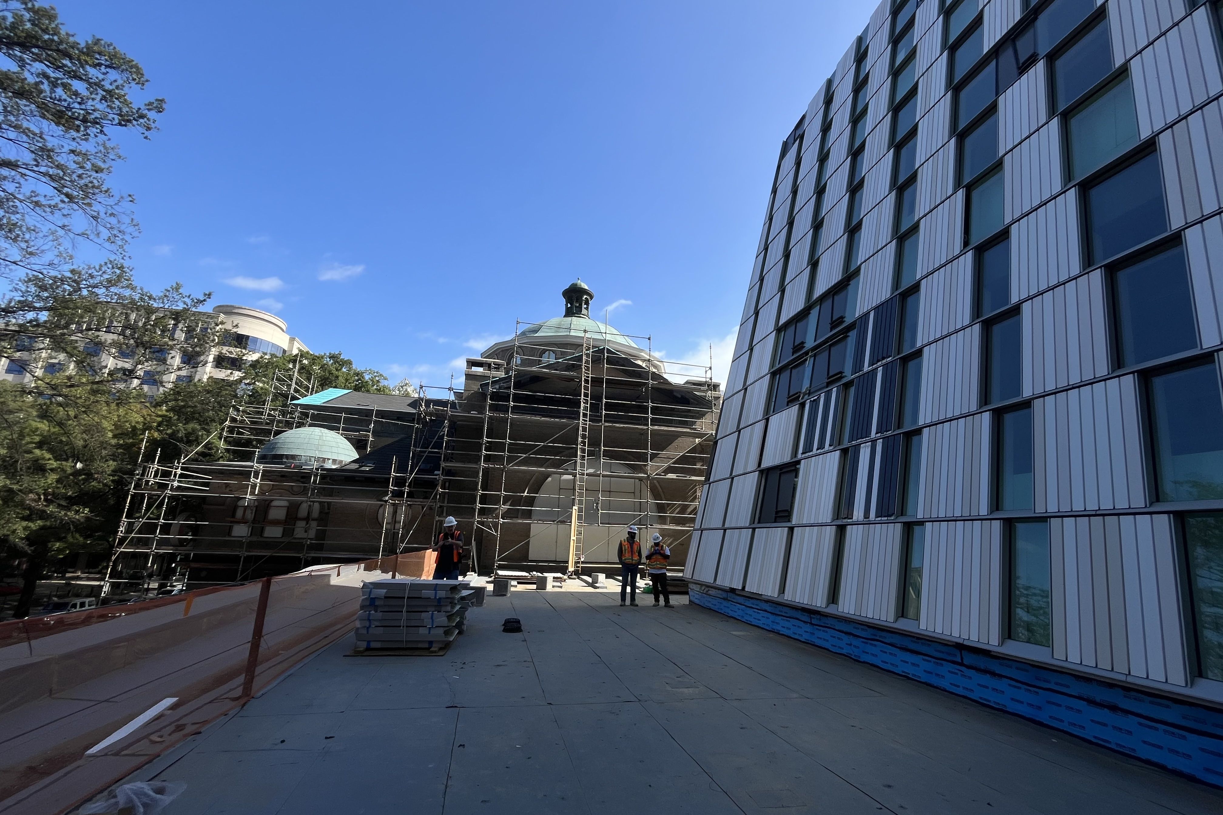 Construction site with workers in orange vests and helmets, scaffolding around a dome-roofed building, modern glass-paneled building on the right, and clear blue sky overhead.