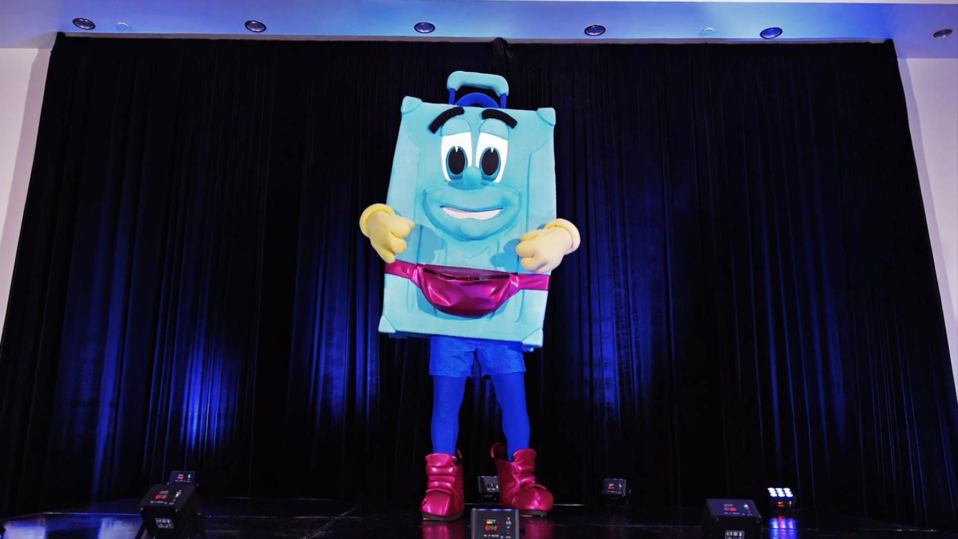 Costumed performer in a large blue cartoon character suit with big eyes and a smile stands on a stage in front of dark curtains, wearing yellow gloves, blue pants, and bright pink boots.