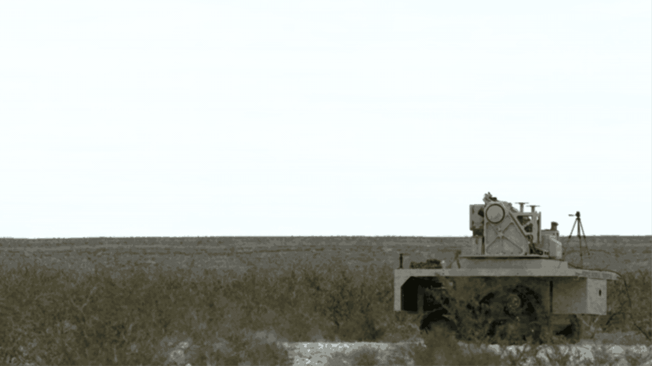A gray, tracked vehicle on rough terrain next to a tripod-mounted camera or sensor, set against a flat, barren landscape under a pale sky.