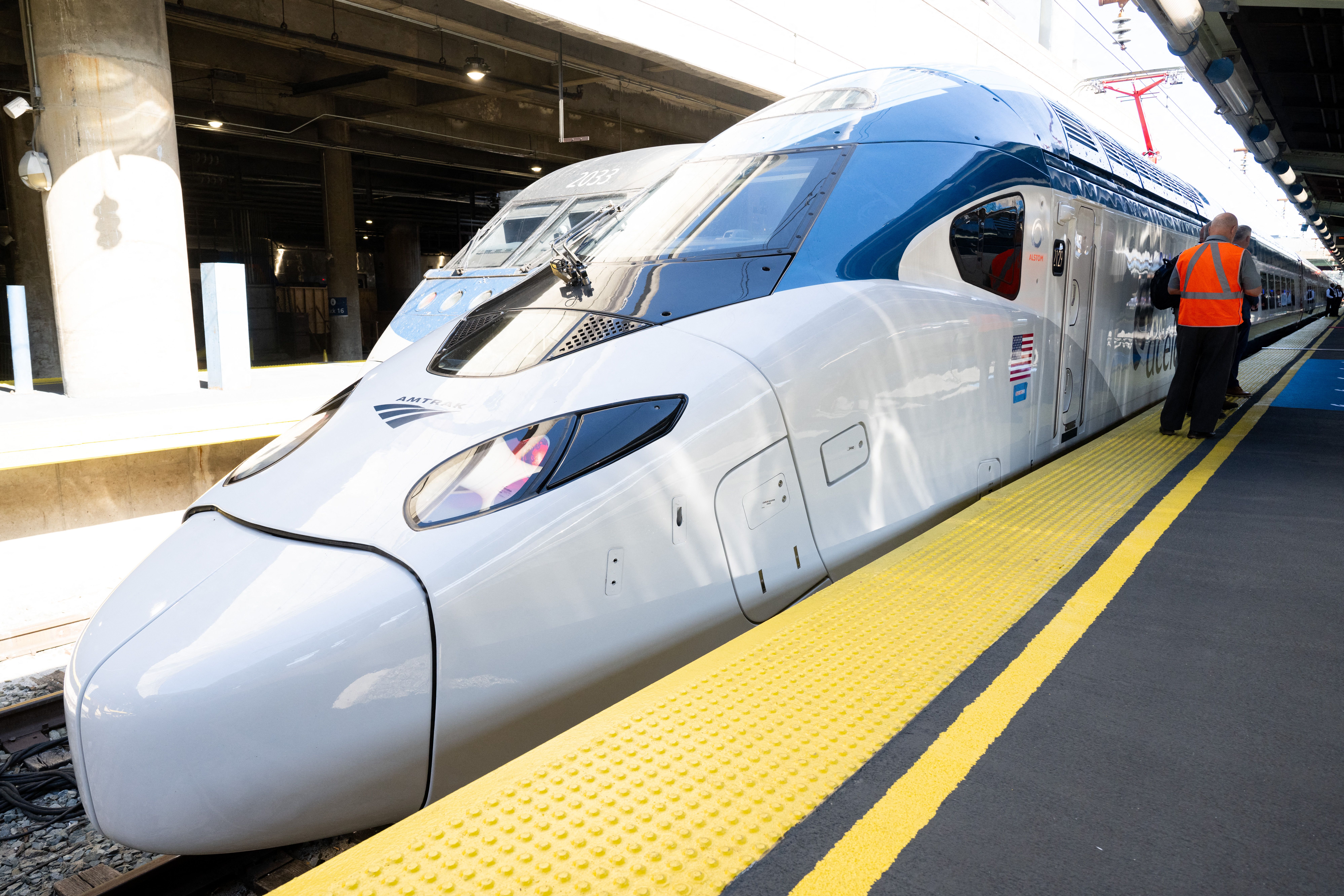 White and blue Amtrak high-speed train at a station platform with yellow safety lines. Two men in orange safety vests stand beside the train under a concrete structure.
