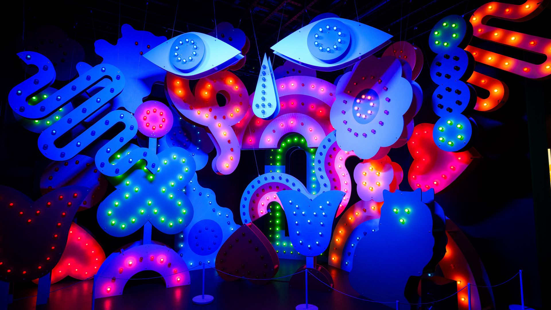 Colorful illuminated abstract shapes including eyes, flowers, hearts, and swirls in blue, pink, red, green, and orange lights suspended in a dark space.
