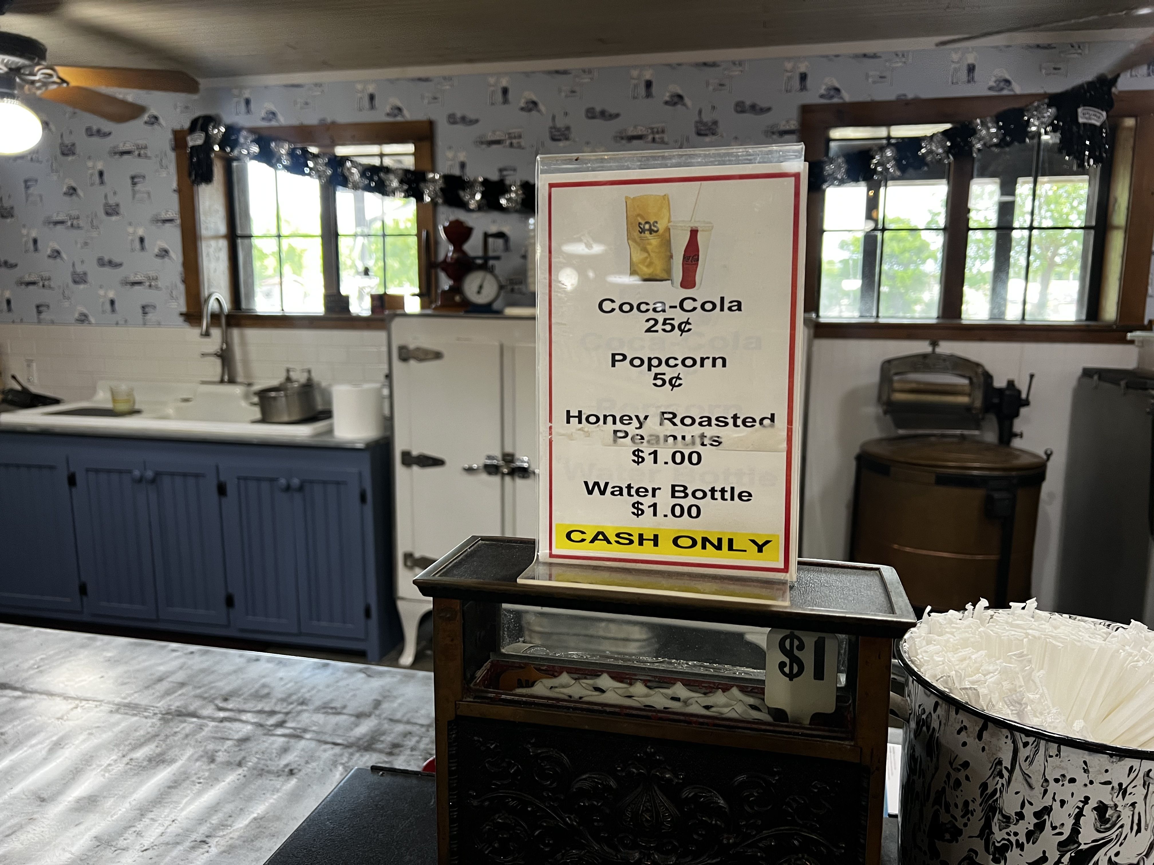 Kitchen scene with blue lower cabinets, white tile counters, and a tall sign listing snacks: Coca-Cola 25¢, Popcorn 5¢, Honey Roasted Peanuts $1.00, Water Bottle $1.00, Cash Only.