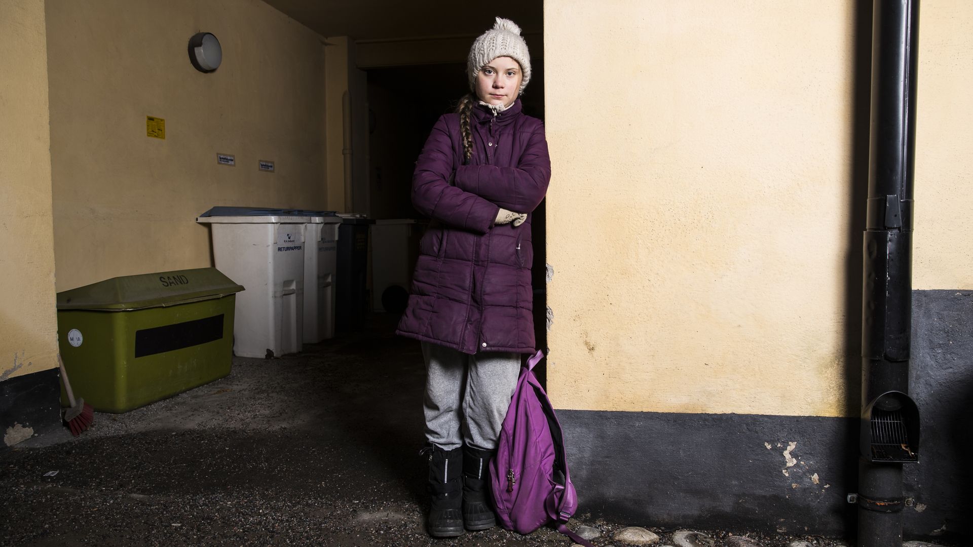 In this image, Greta stands in a puffy winter coat and hat while leaning against a wall, with a bookbag near her.