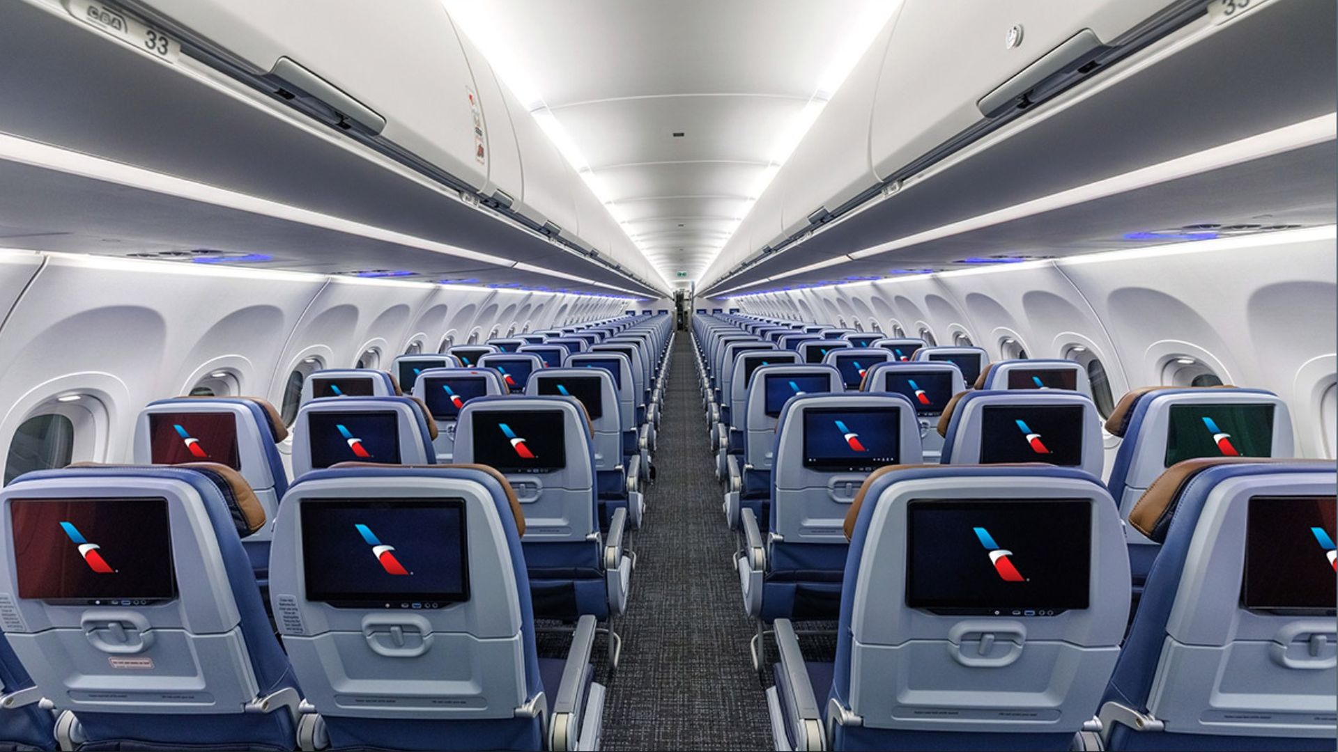 Inside a modern airplane cabin with a long central aisle, rows of blue seats each with a personal screen on the back, bright white overhead panels, and blue accent lighting along the cabin sides.