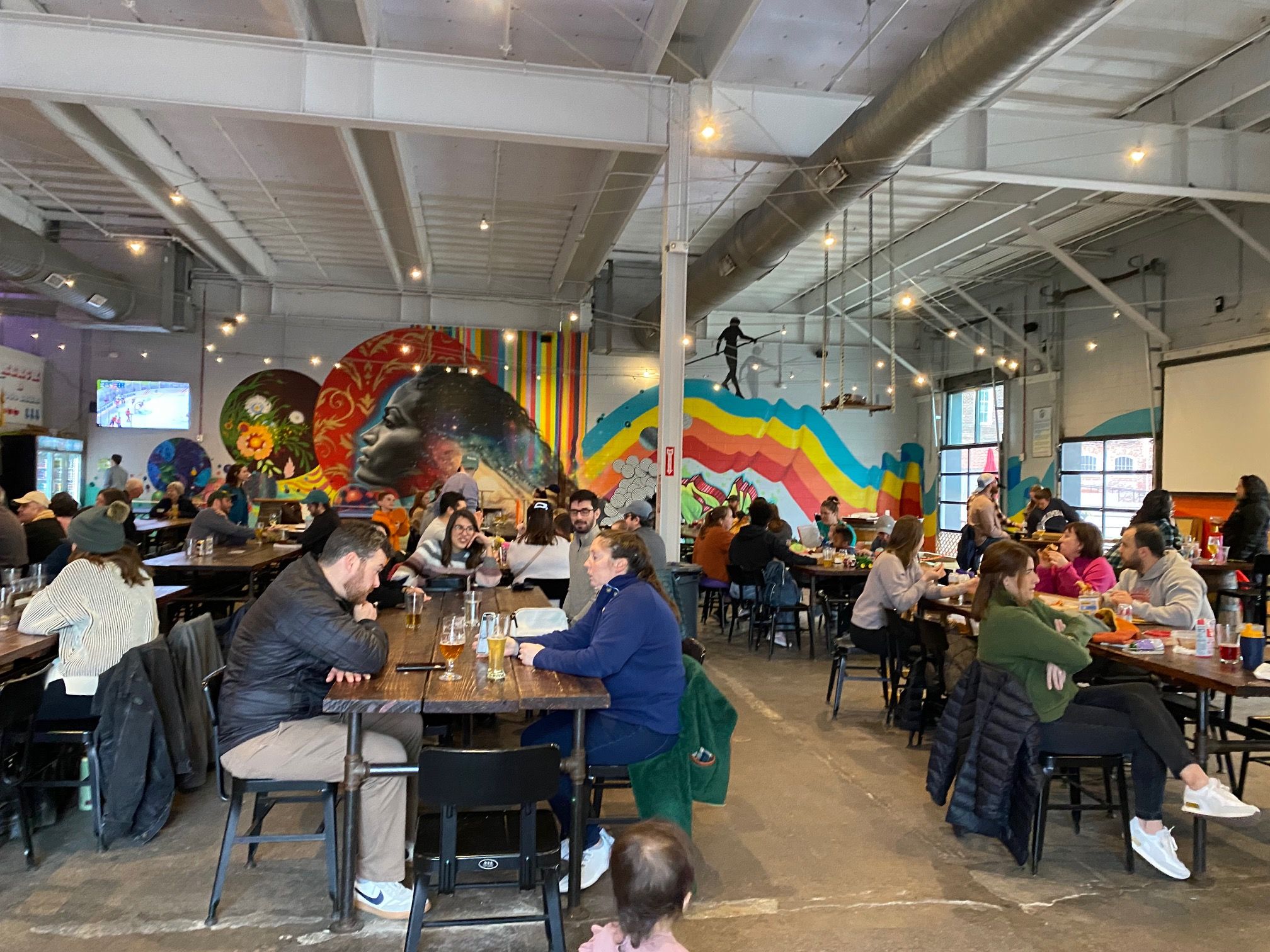 Busy indoor space with people sitting at wooden tables, colorful murals on the walls including a profile of a woman's face and rainbow stripes, string lights overhead, and large windows.