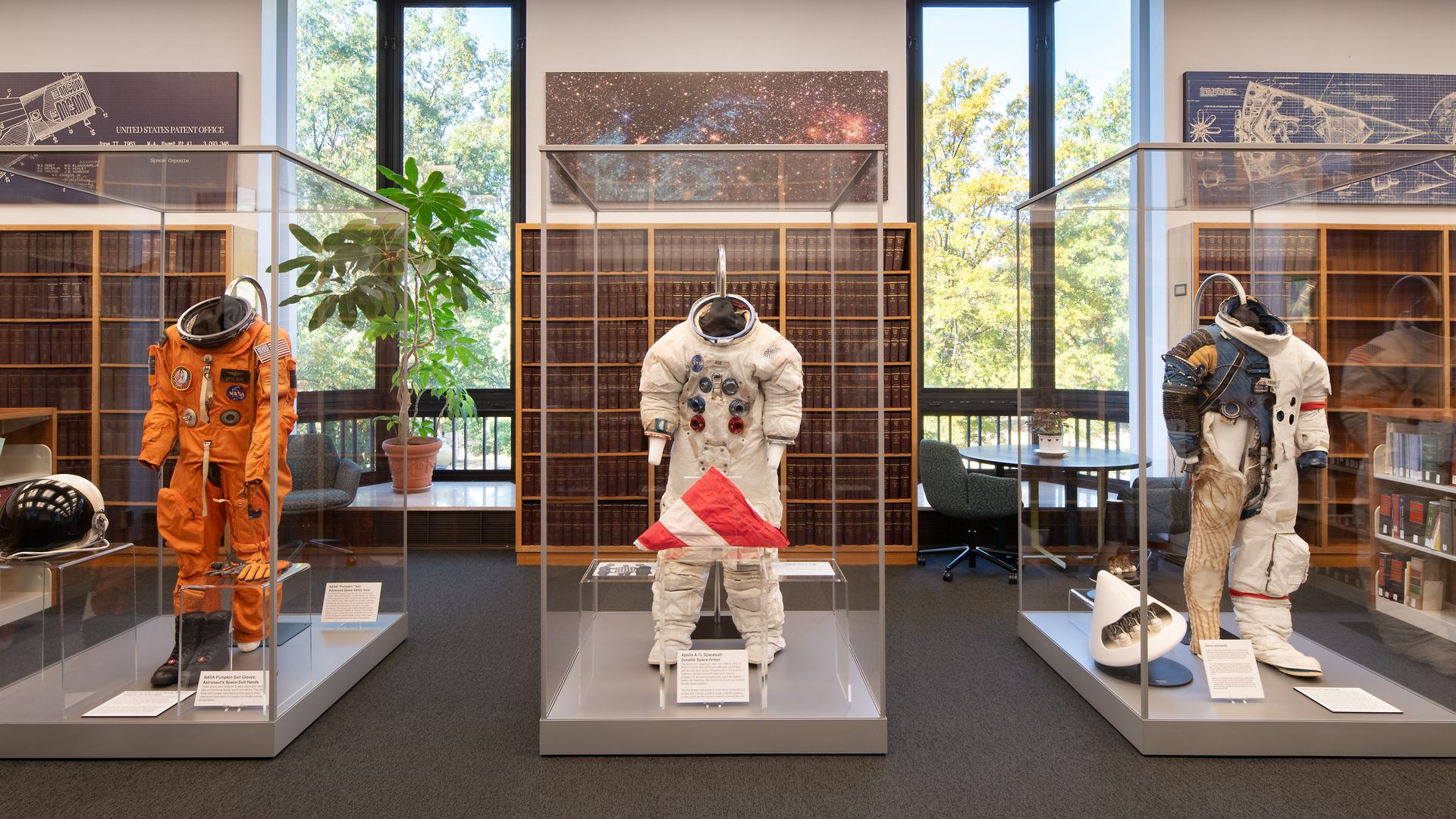 Exhibit of three spacesuits in glass cases: orange left, white center, and white right with patches. Behind are tall bookshelves, large windows with trees, and space-themed wall art.