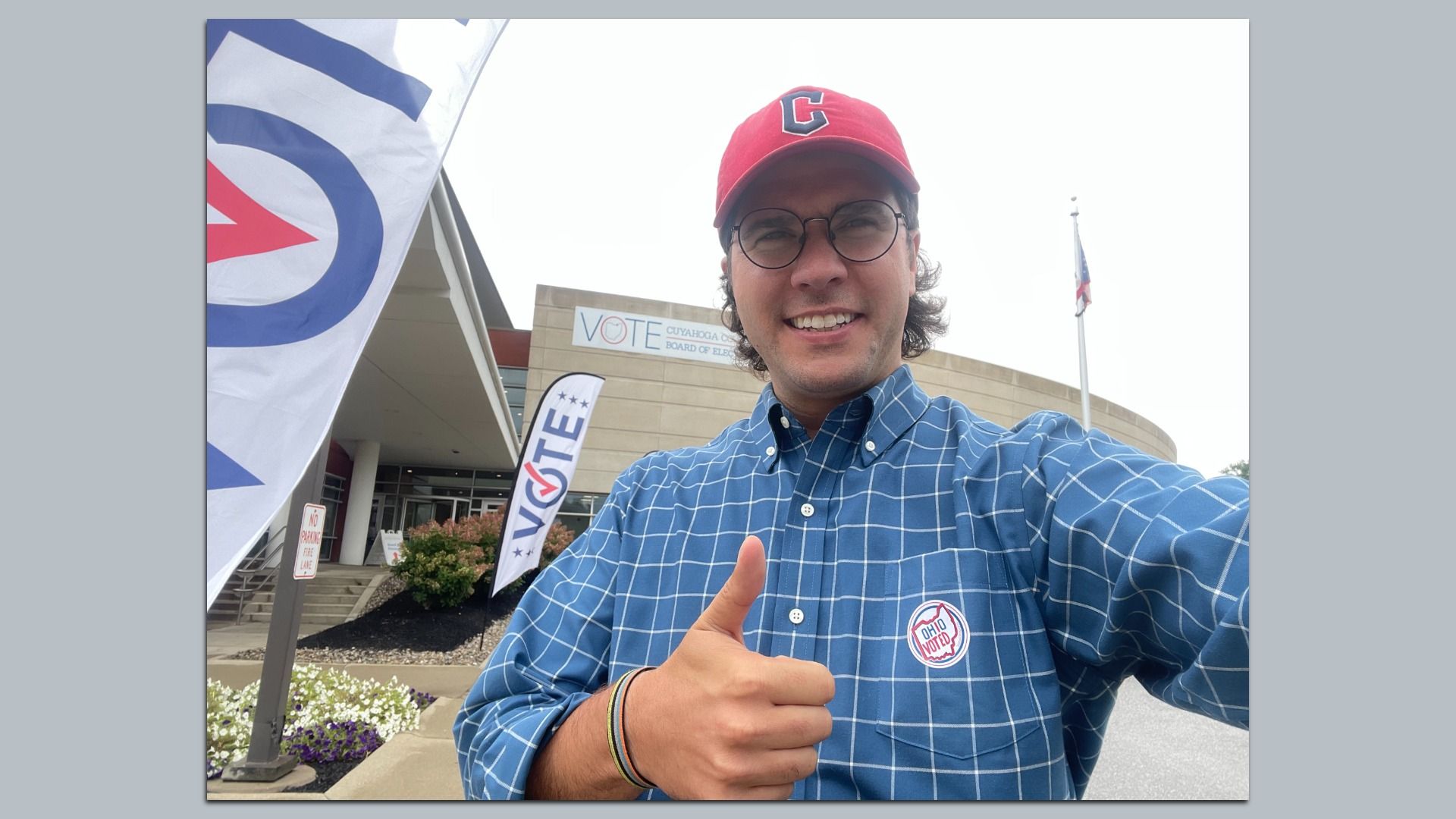 Man wearing red hat and blue checkered shirt with an "Ohio Voted" sticker, giving thumbs up outside a voting center with "VOTE" flags and a building sign for Cuyahoga County Board of Elections.