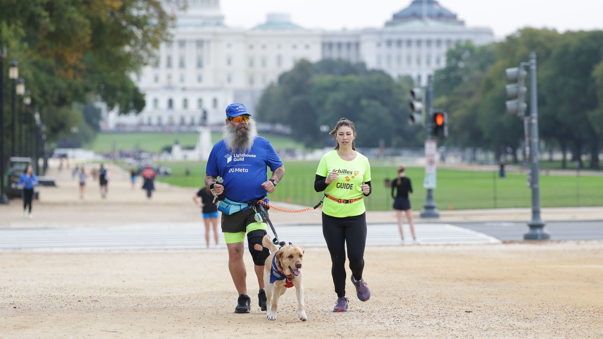 Two runners in the foreground, a man in blue and a woman in neon, run with a leashed dog on a gravel path. In the distance, the U.S. Capitol dome and another building appear behind trees.