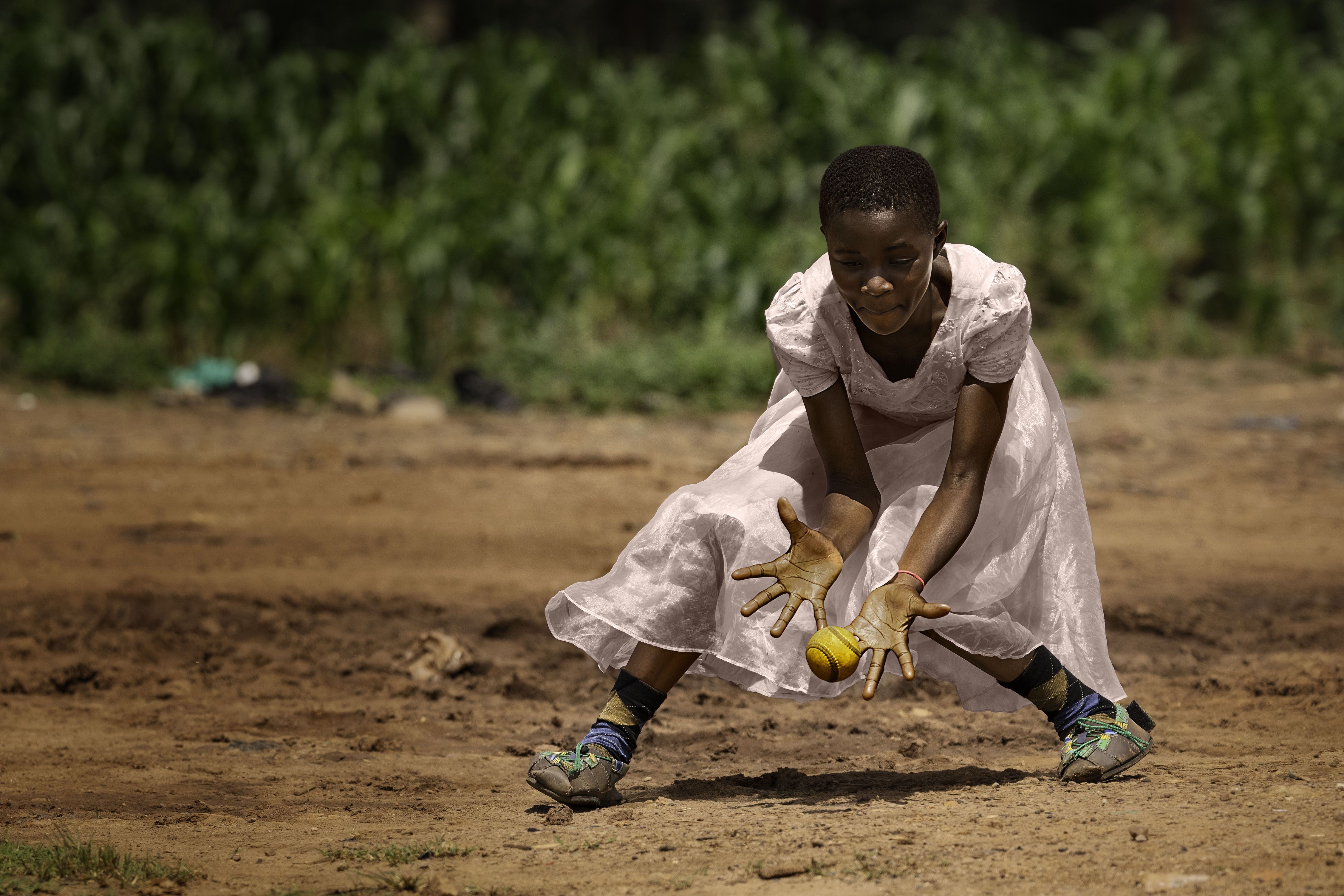 A young girl in a white dress crouches on a dusty field, hands stretched toward a yellow ball. She wears colorful sneakers, with green foliage blurred in the background.