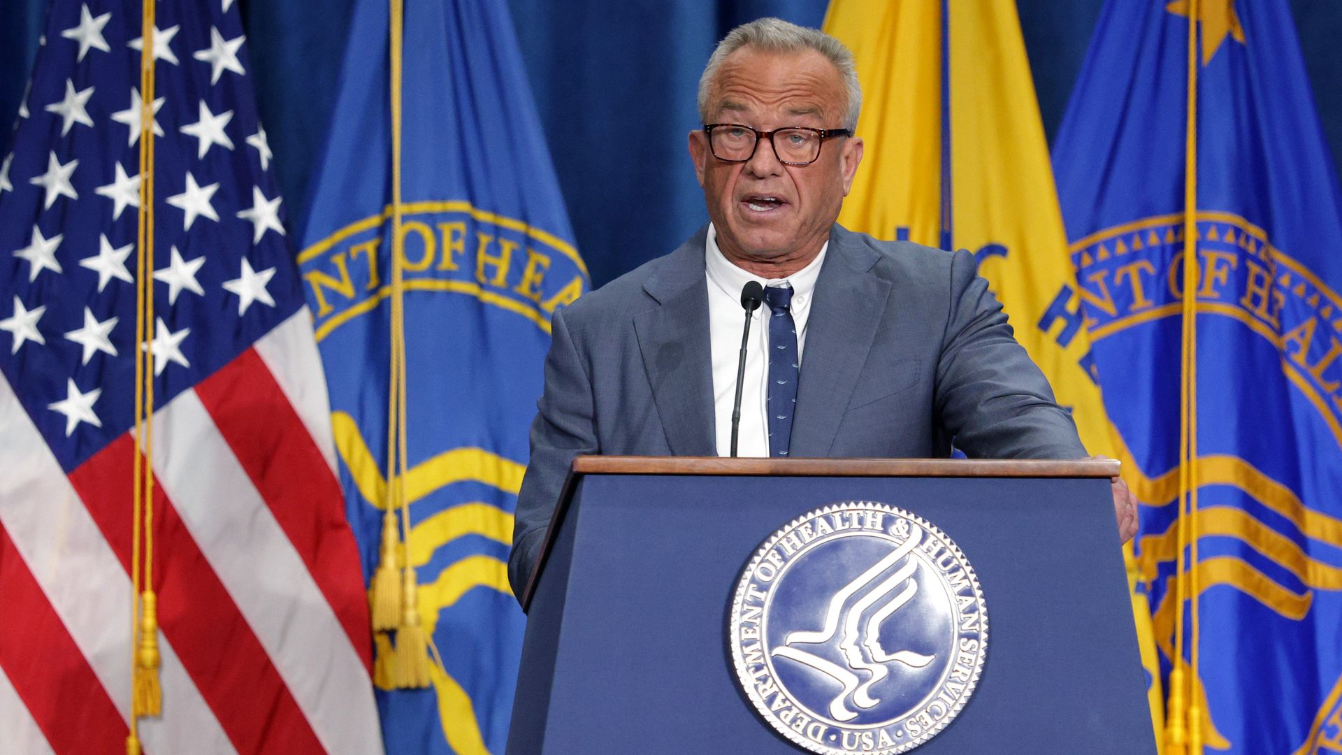 A fair-skinned, gray-haired, bespectacled Health and Human Services Secretary Robert F. Kennedy Jr., wearing a gray suit jacket, white shirt and blue tie, speaks at a blue podium.