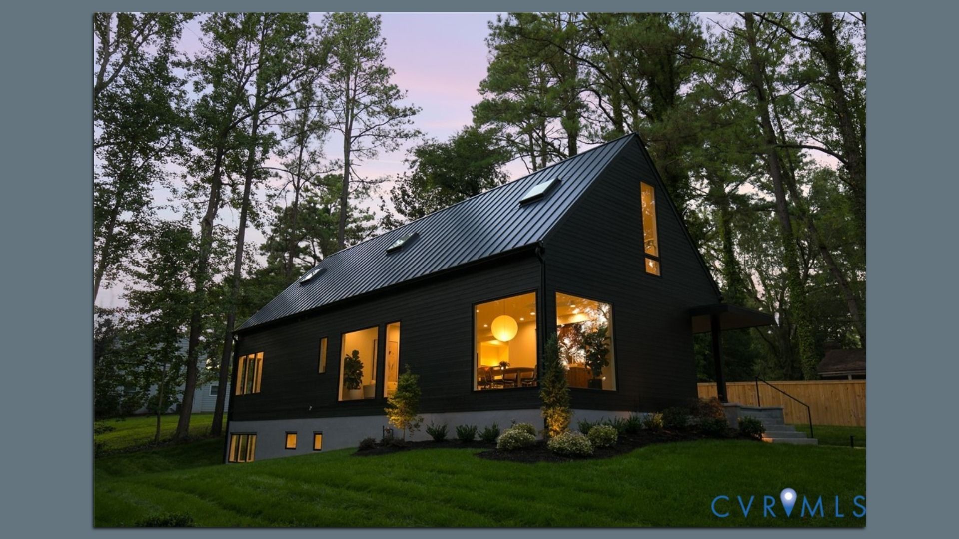 Modern black two-story house with a sloped metal roof and large windows glowing warm inside, set among tall trees on a green lawn at dusk; a wooden fence and stairs are visible on the right.